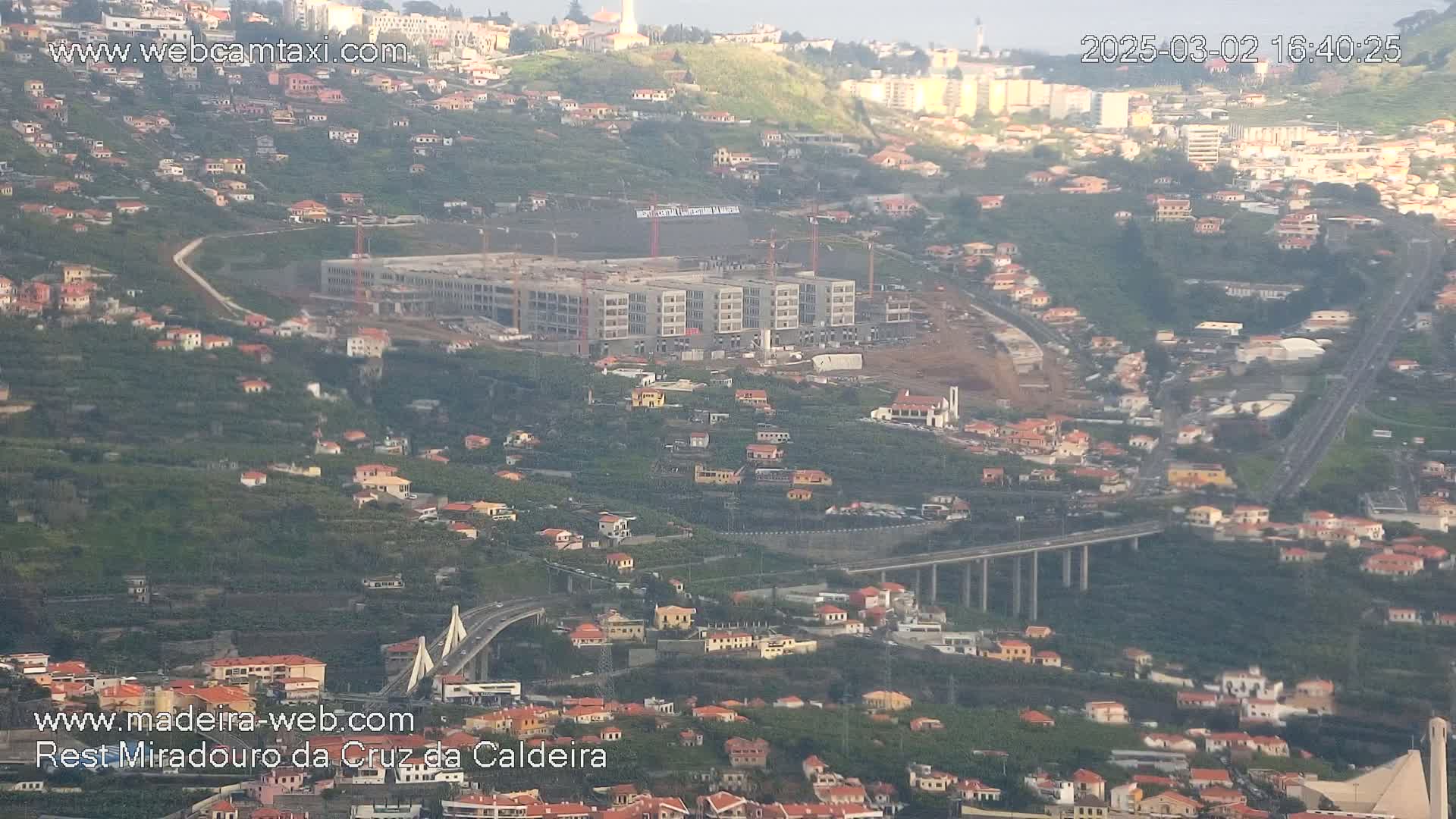 An aerial view shows a hillside town with a large building under construction, a long elevated highway, and a cable-stayed bridge, all under hazy, sunny conditions.