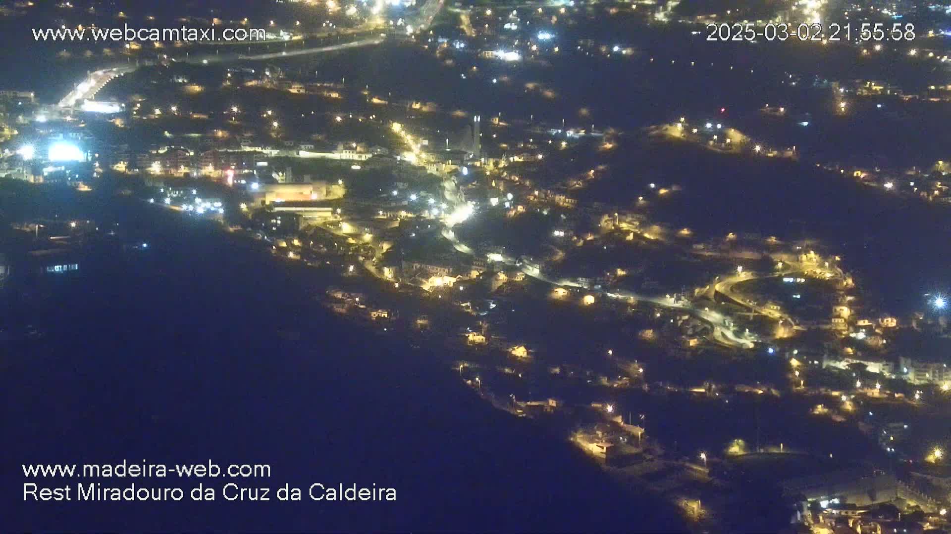 A nighttime, high-angle view shows a town nestled on a hillside, illuminated by numerous lights.