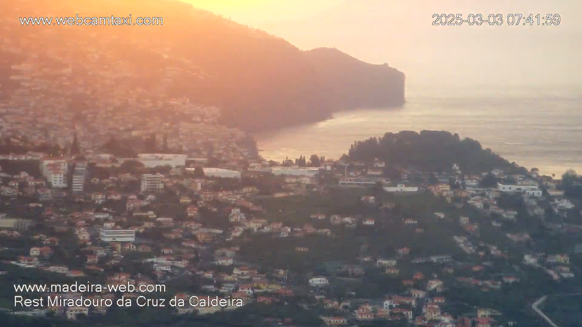 A coastal town nestled on hillsides overlooking a calm ocean at sunrise, bathed in warm, orange light.
