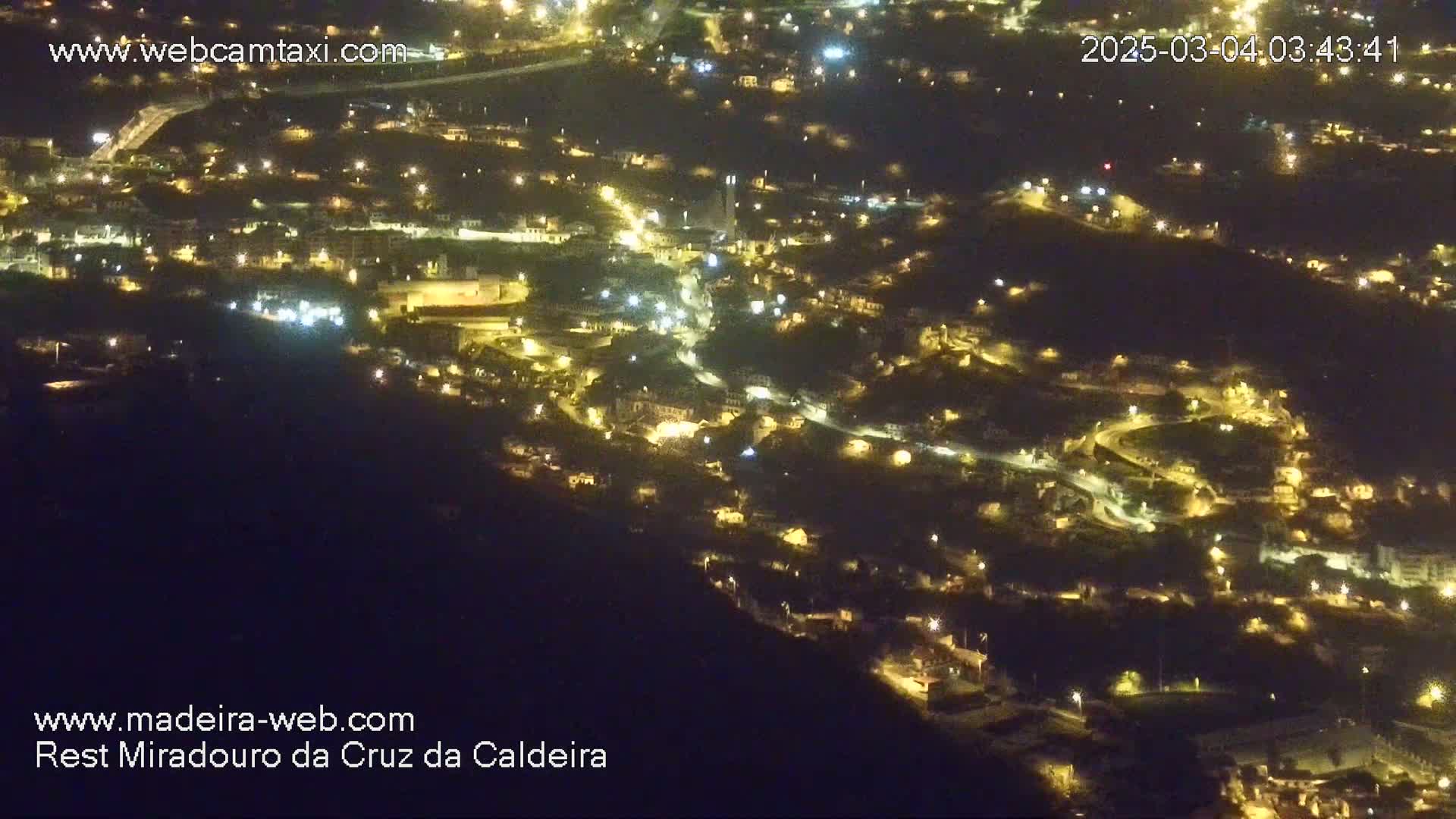 A nighttime aerial view shows a hillside town illuminated by numerous lights.
