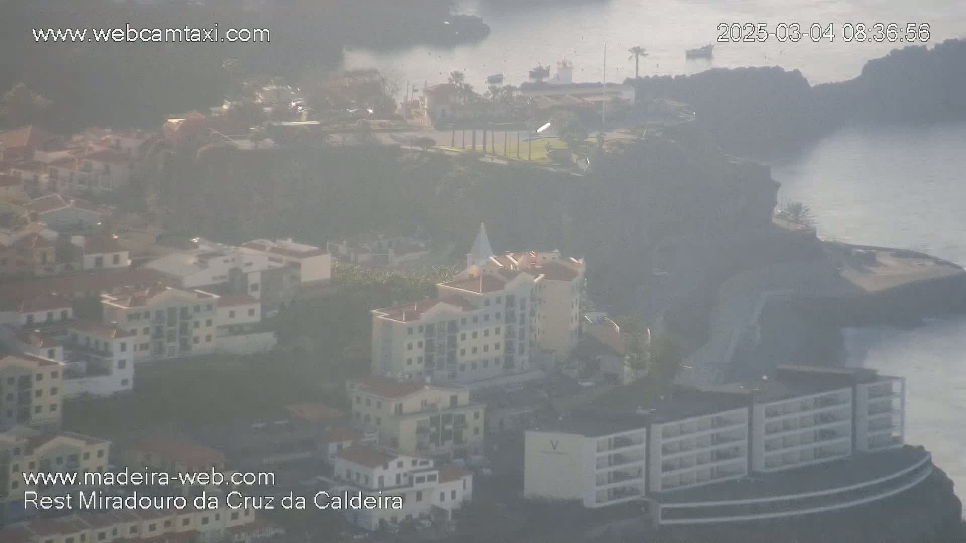A hazy coastal town with multi-story buildings, cliffs, and a body of water is seen from an aerial perspective.