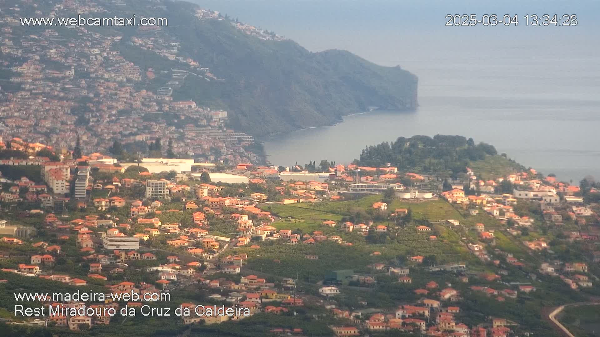 An aerial view shows a coastal town with numerous orange-roofed houses nestled on a hillside overlooking a calm ocean, under a hazy sky.