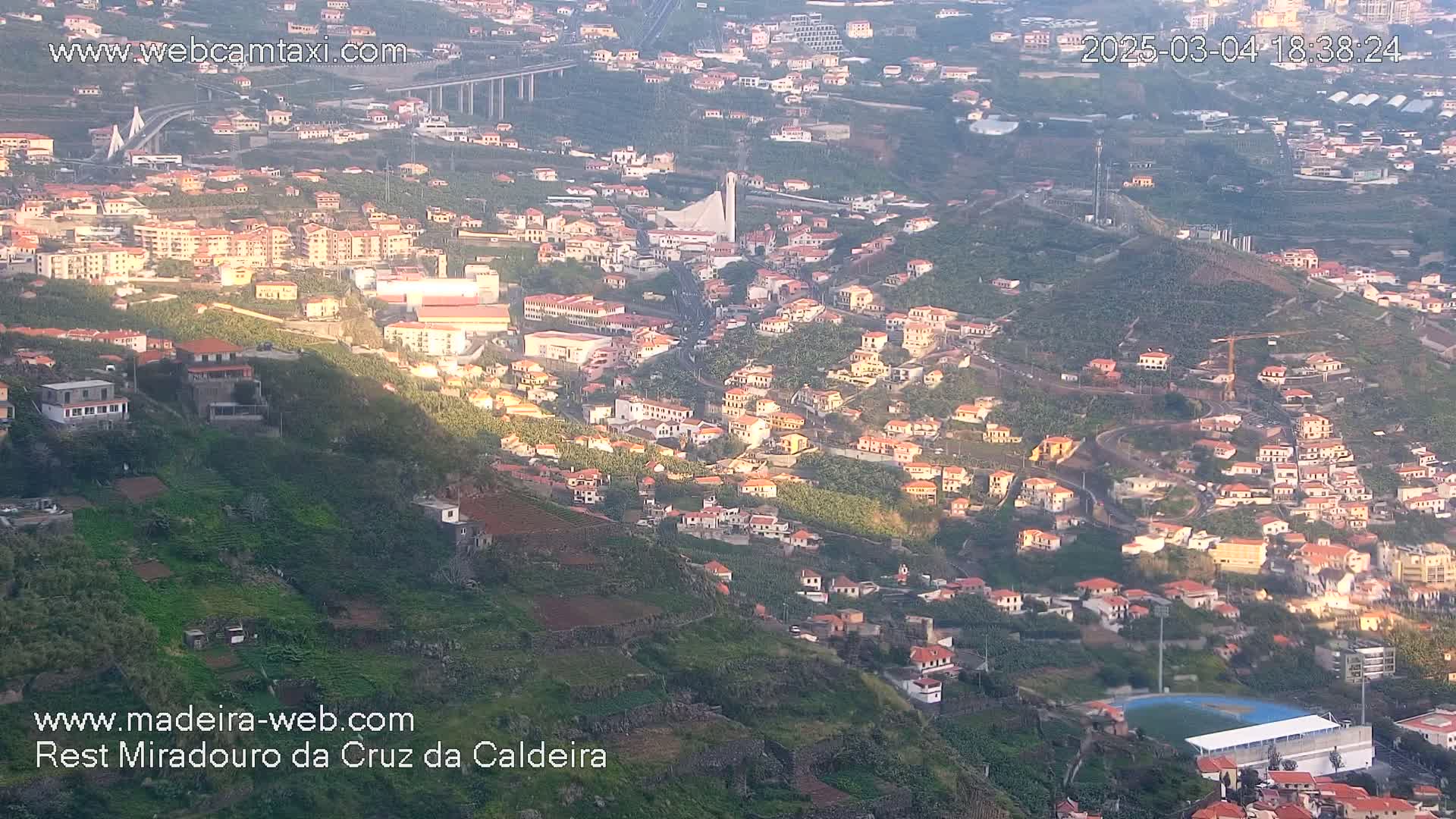 A hillside town with red-roofed houses and terraced farmland is bathed in the soft light of late afternoon or early evening.