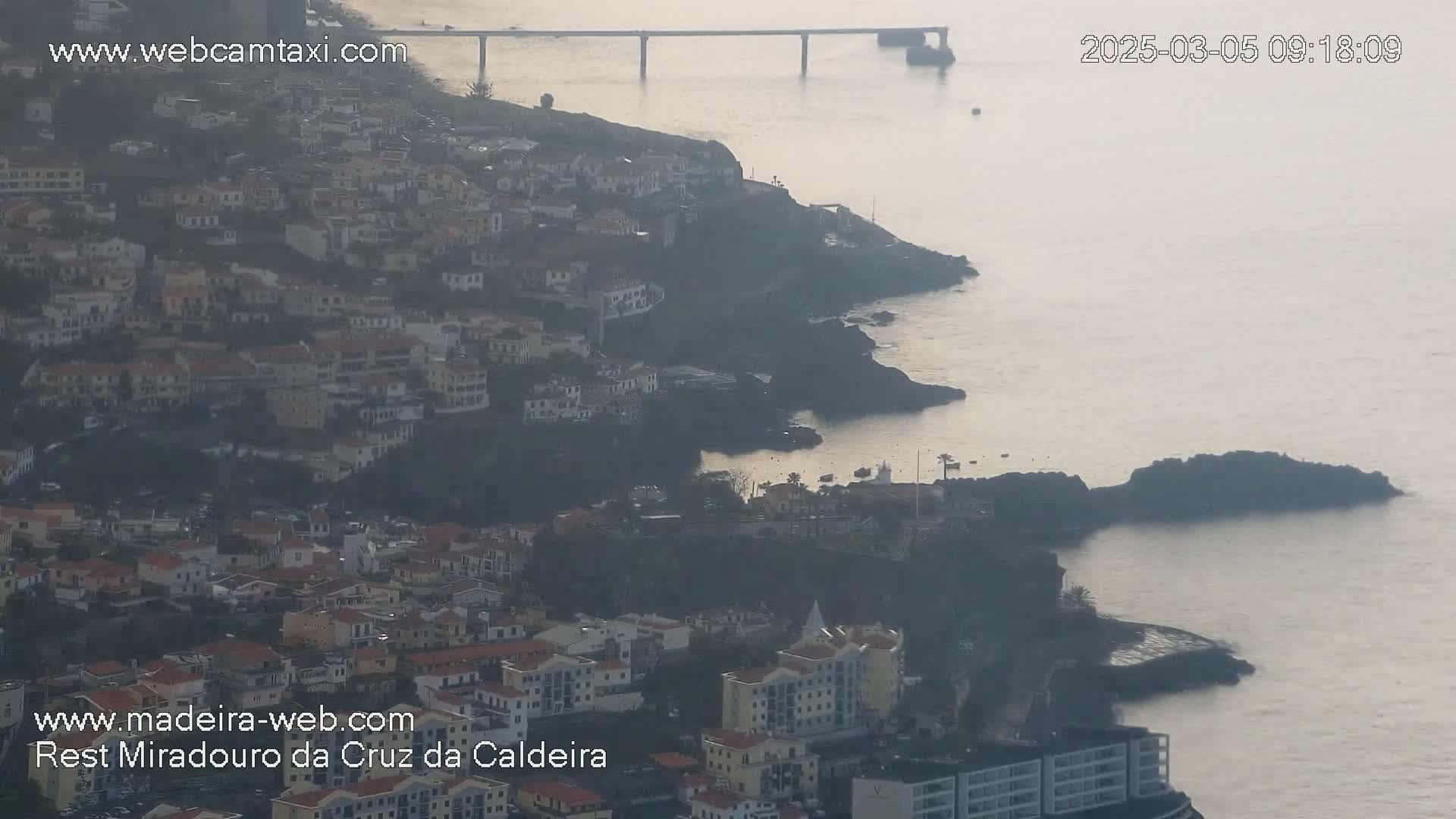 A hazy coastal town clings to a rocky hillside, overlooking a calm sea and a distant bridge under a muted sky.
