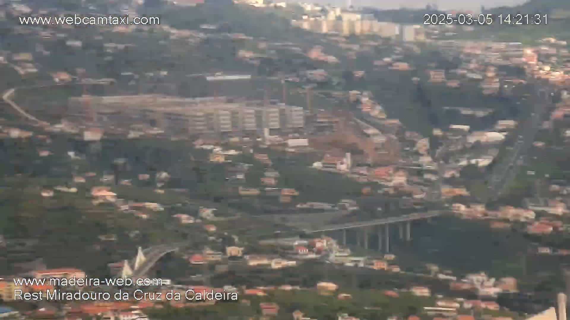 A hazy, high-angle view shows a sprawling city nestled in a hillside, featuring a large bridge and a partially visible construction site.
