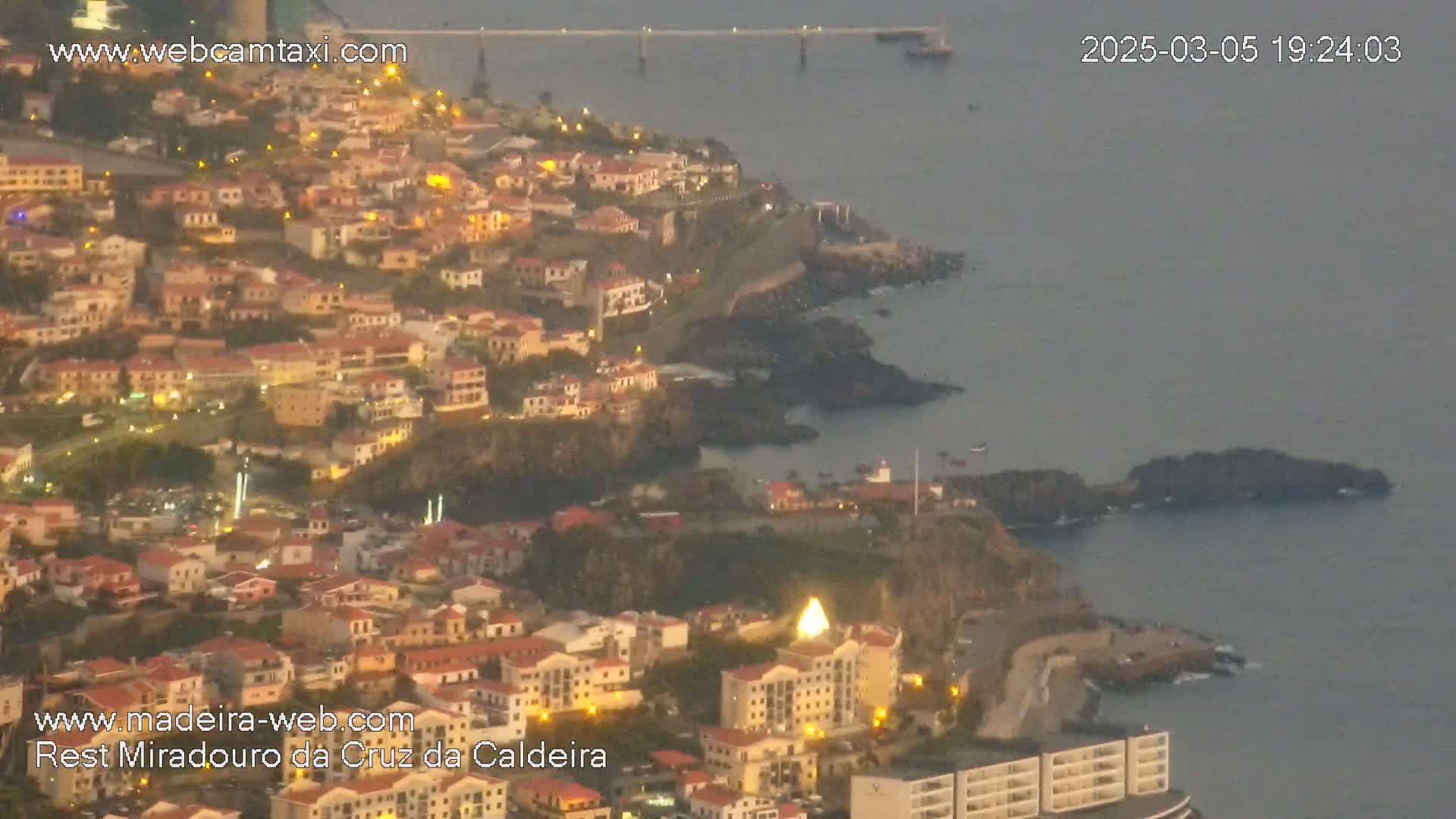 A coastal town with many lights is nestled along a dark rocky shoreline, overlooking a calm body of water at dusk.