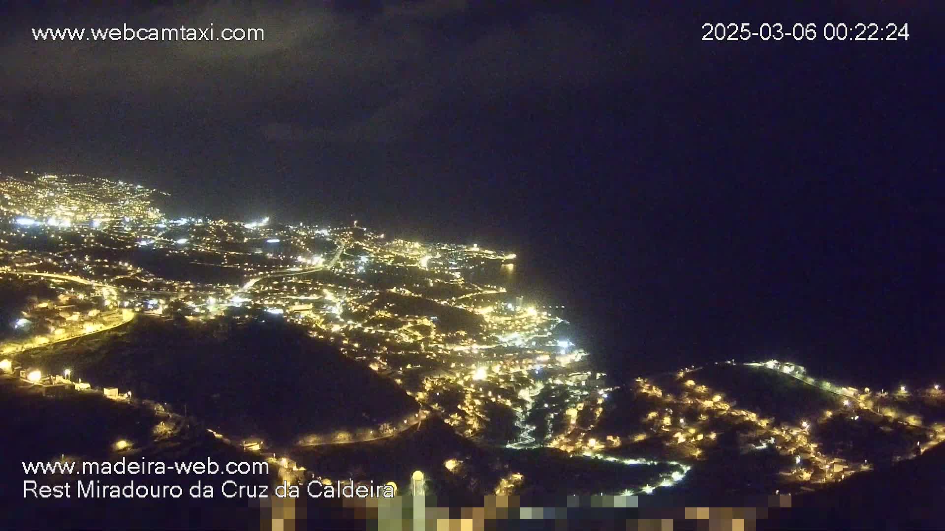 A nighttime, aerial view shows a coastal city illuminated with many lights, overlooking a dark ocean under a clear, dark sky.