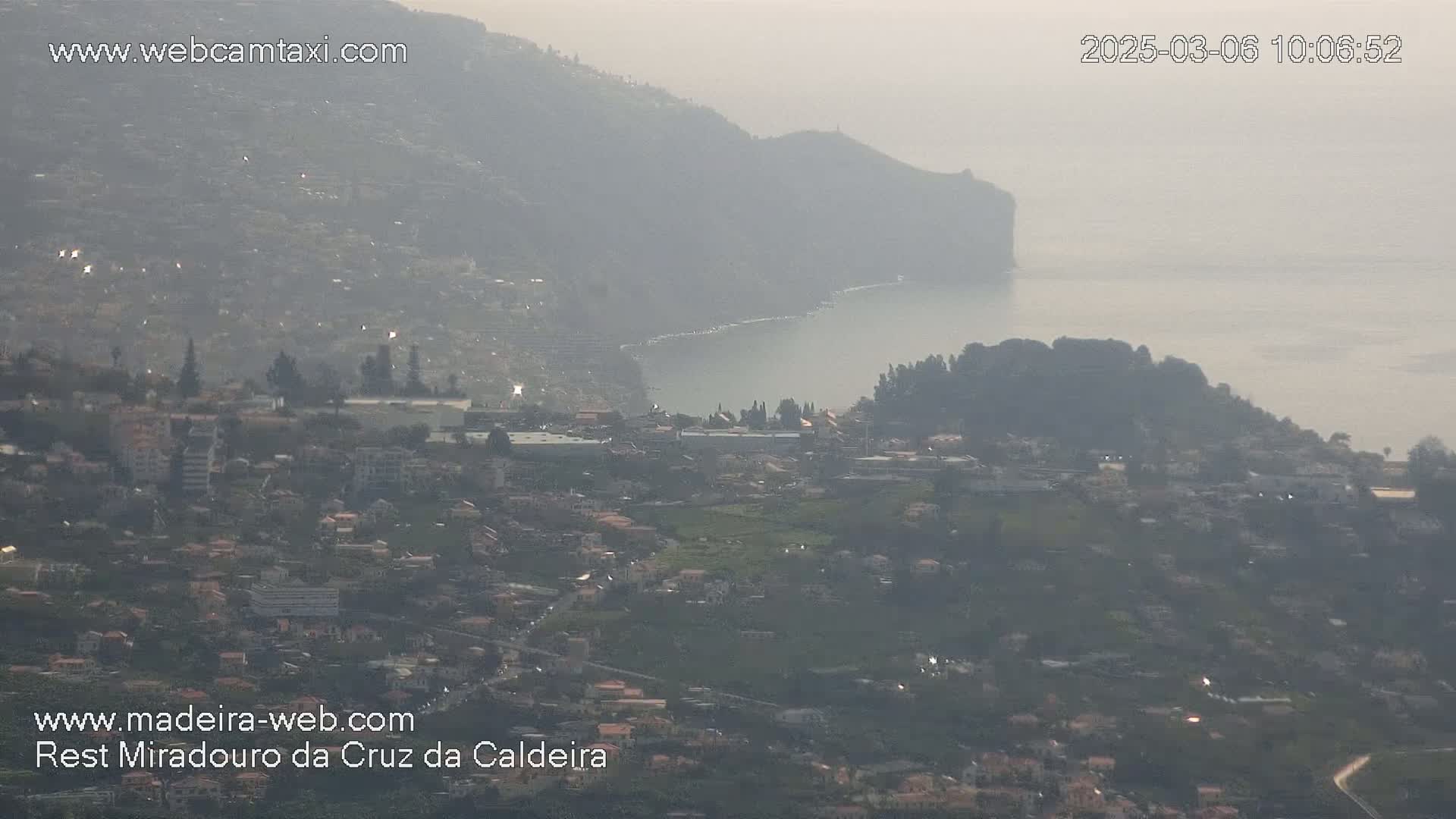 A hazy view shows a coastal town nestled between hills, overlooking a calm body of water.