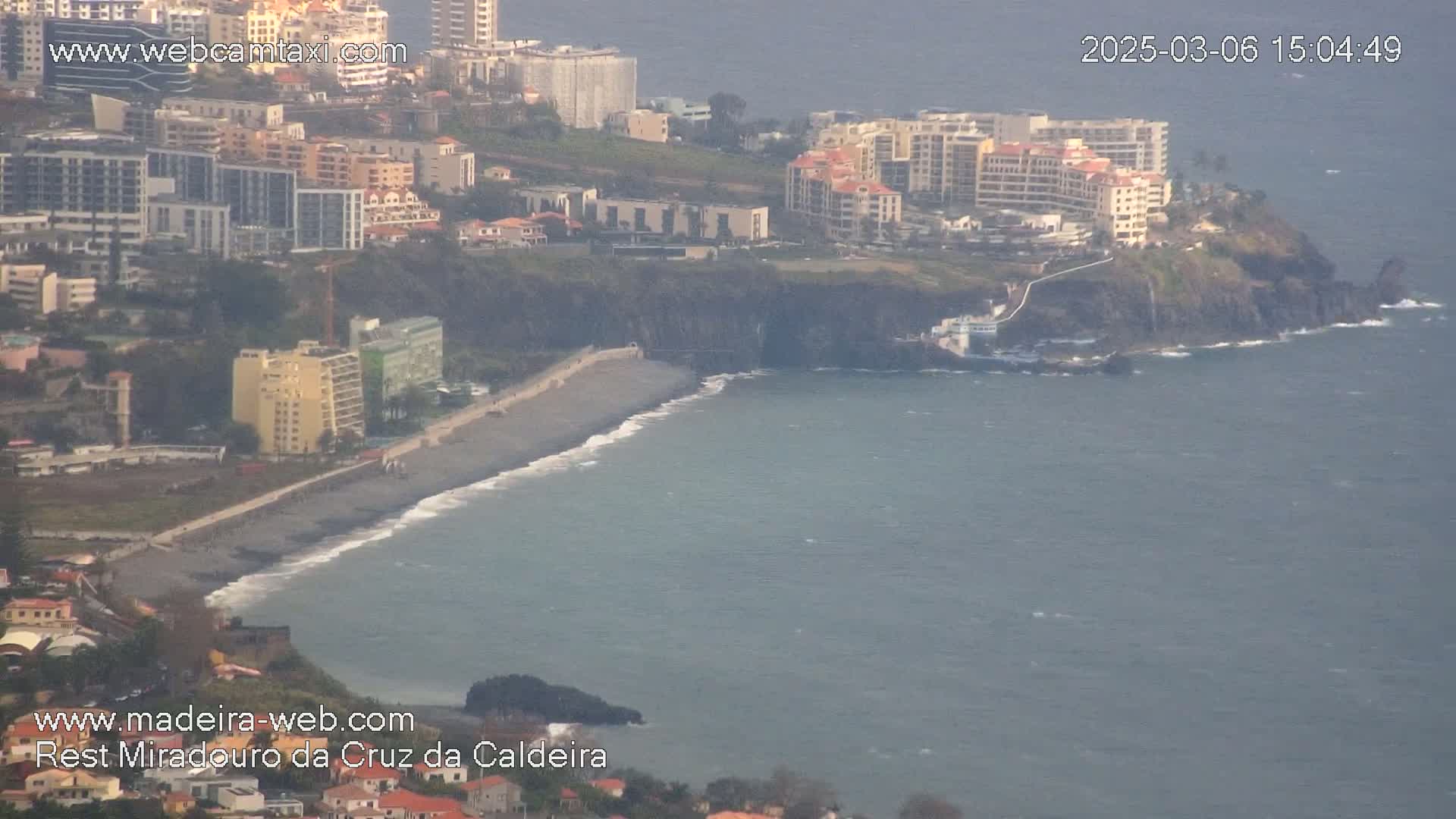 A dark-sand beach curves along a rocky coastline, bordered by multi-story buildings and a slightly hazy ocean under a muted sky.
