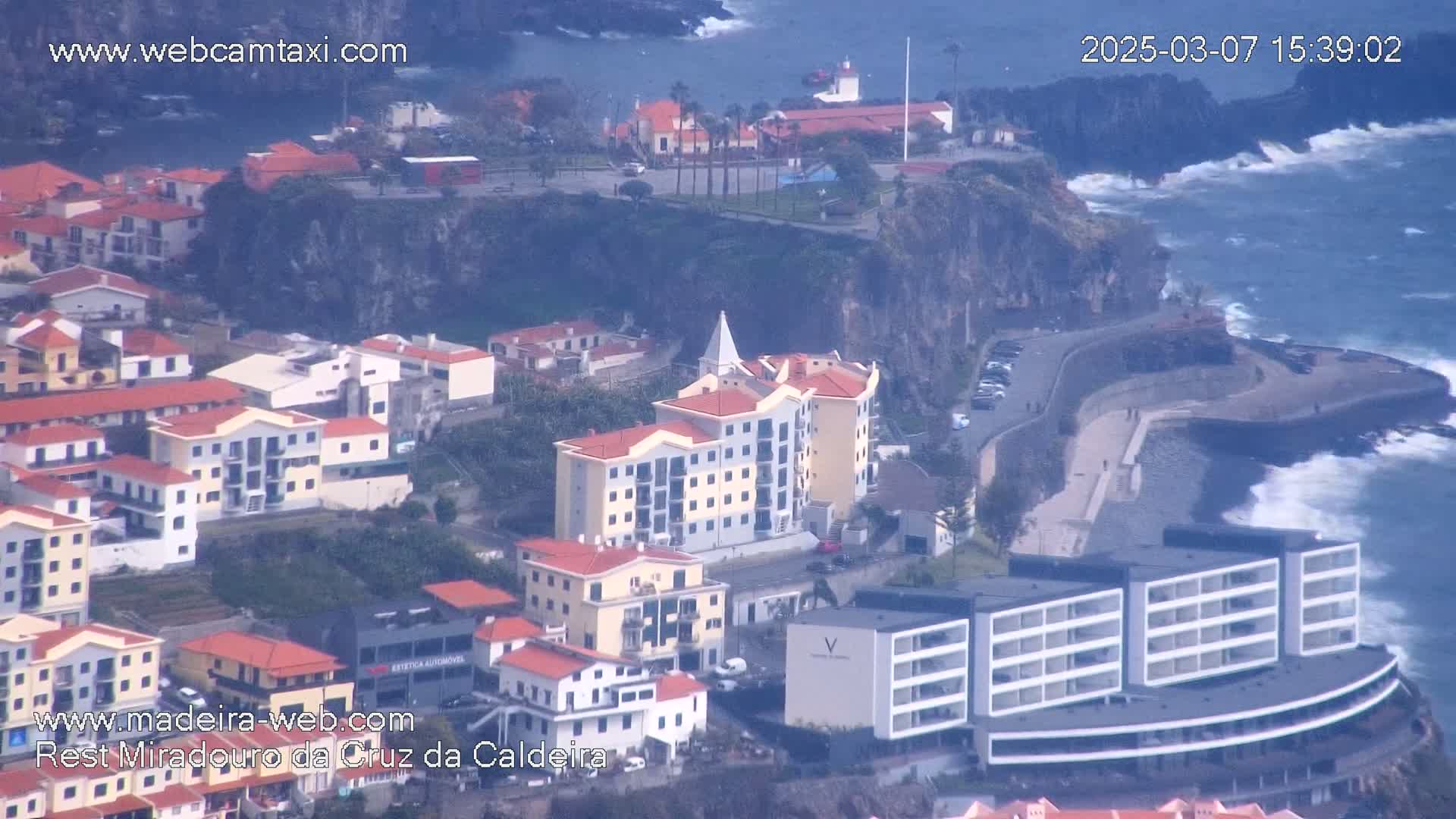 An aerial view shows a coastal town with multi-story buildings, a rocky shoreline, and a choppy ocean under a cloudy sky.