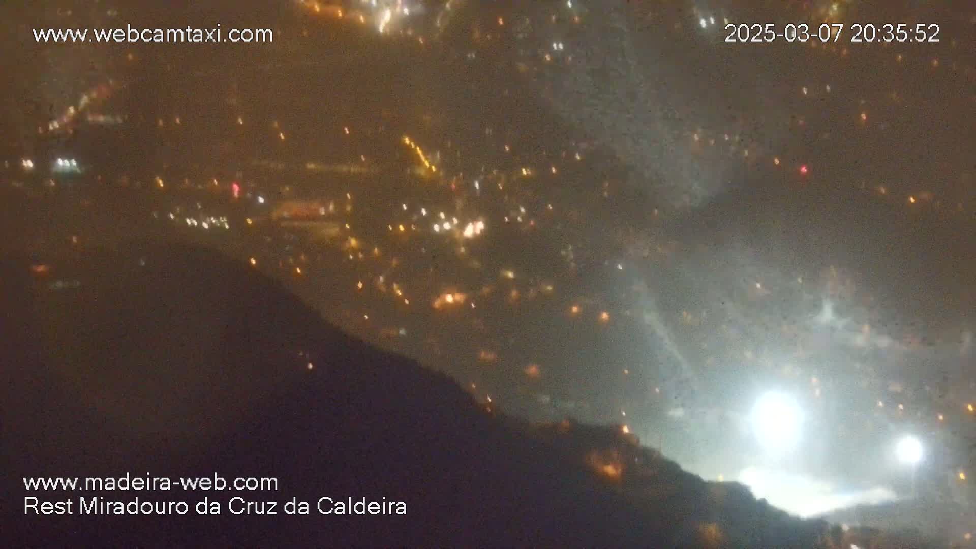 A nighttime, hazy view from a high vantage point shows a distant town speckled with lights below a dark, hazy sky where bright lights are also visible.