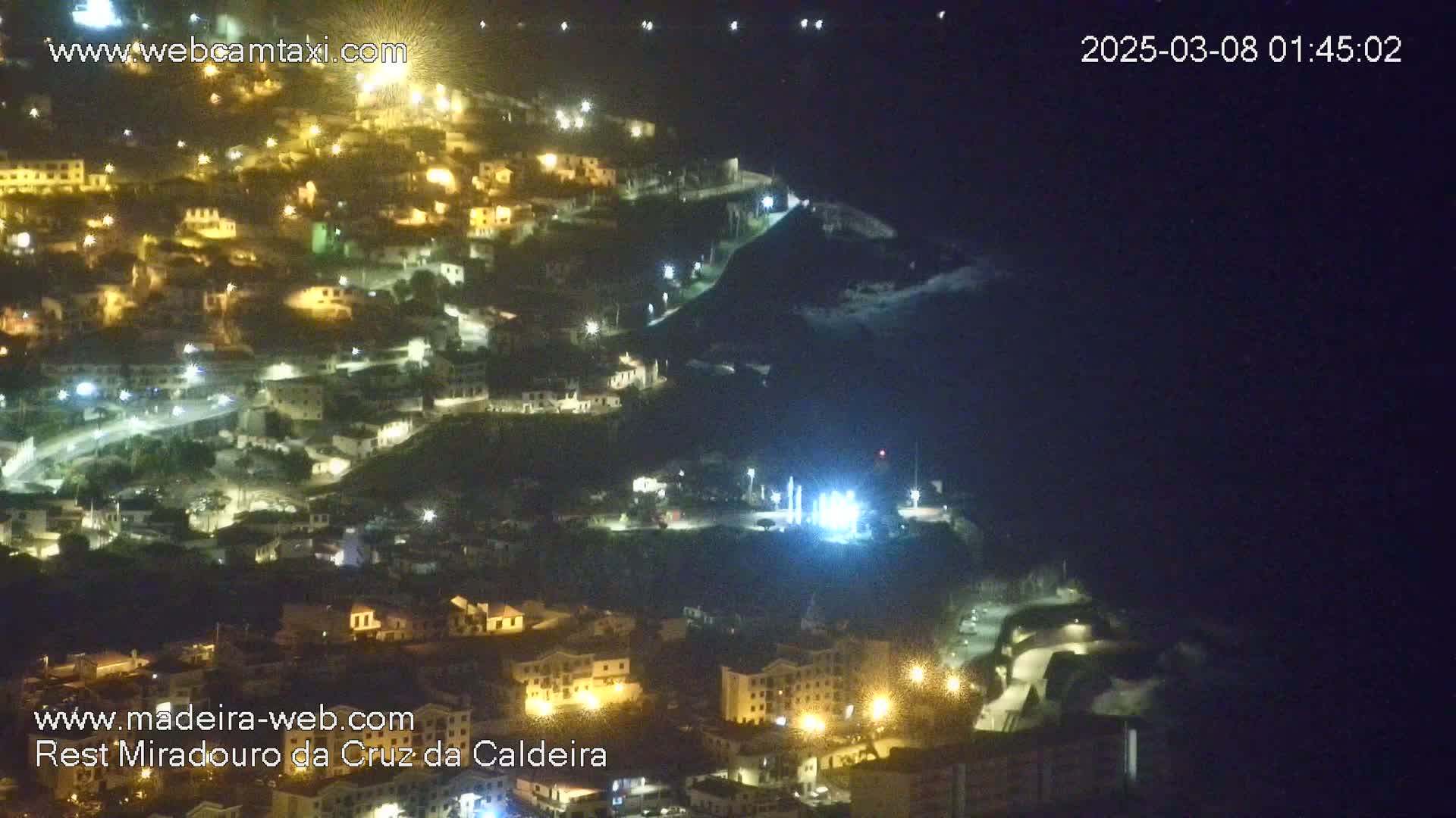 A nighttime, high-angle view of a coastal town illuminated by streetlights, with the ocean visible in the darkness.
