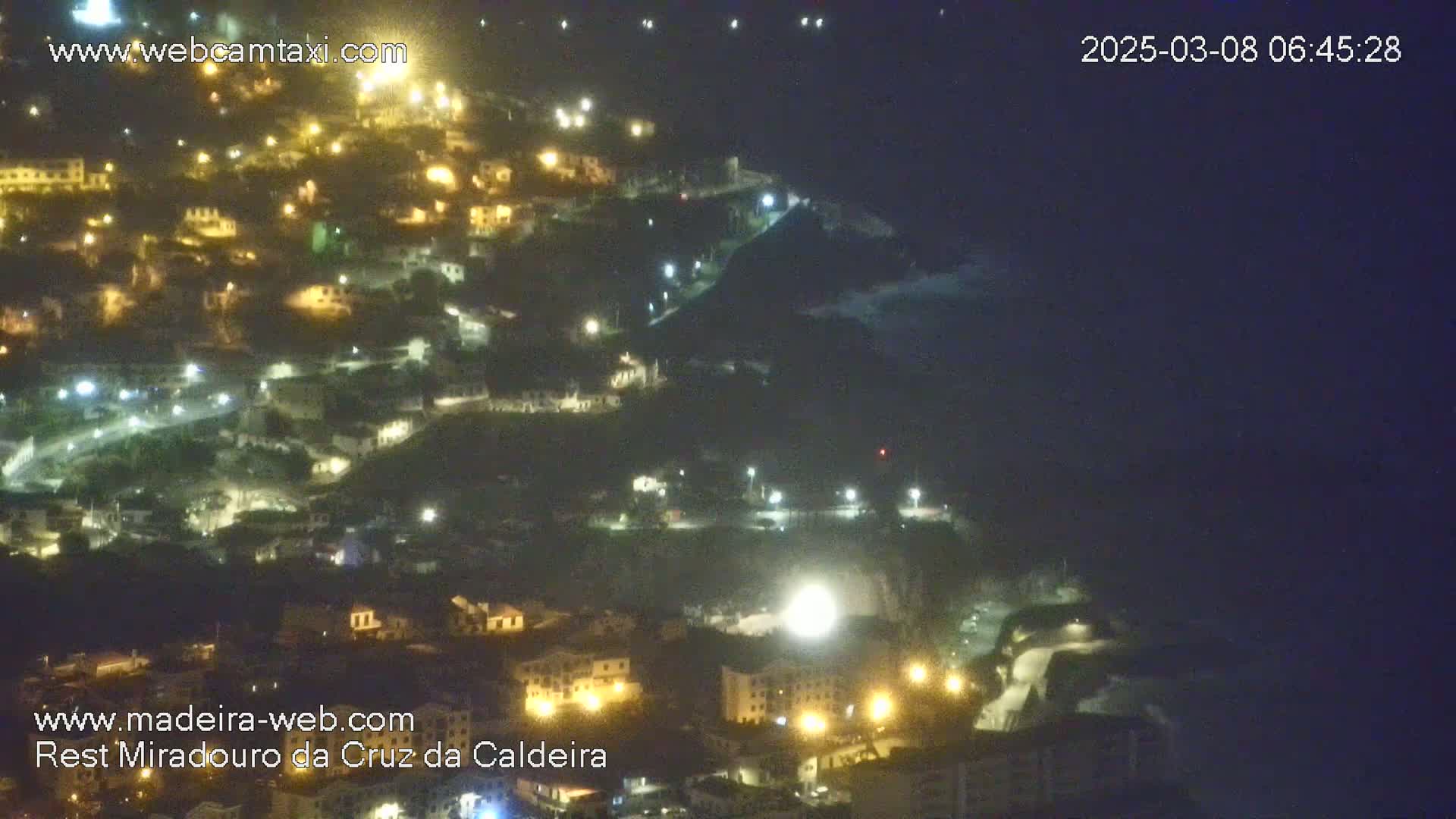 A nighttime, aerial view of a hillside town illuminated by streetlights, overlooking a dark ocean.