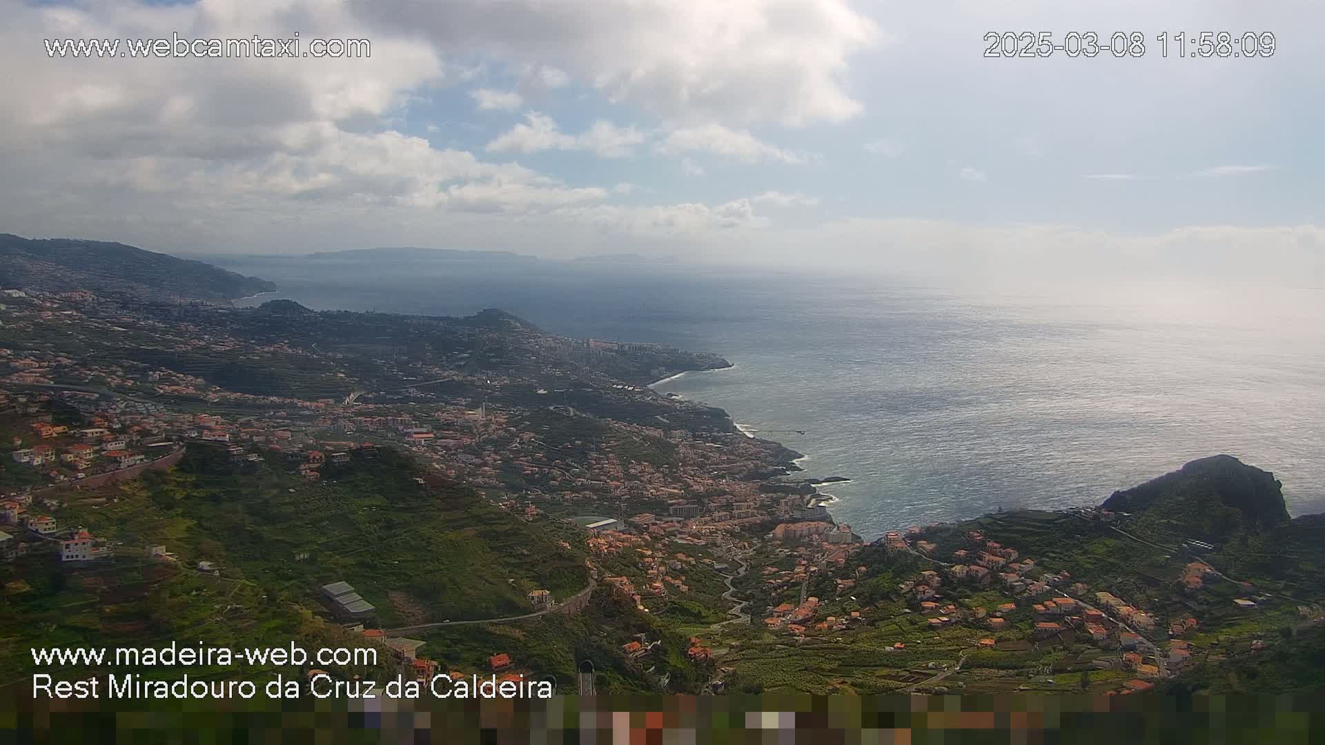 A coastal town nestled into a hillside overlooking a calm ocean under a partly cloudy sky.