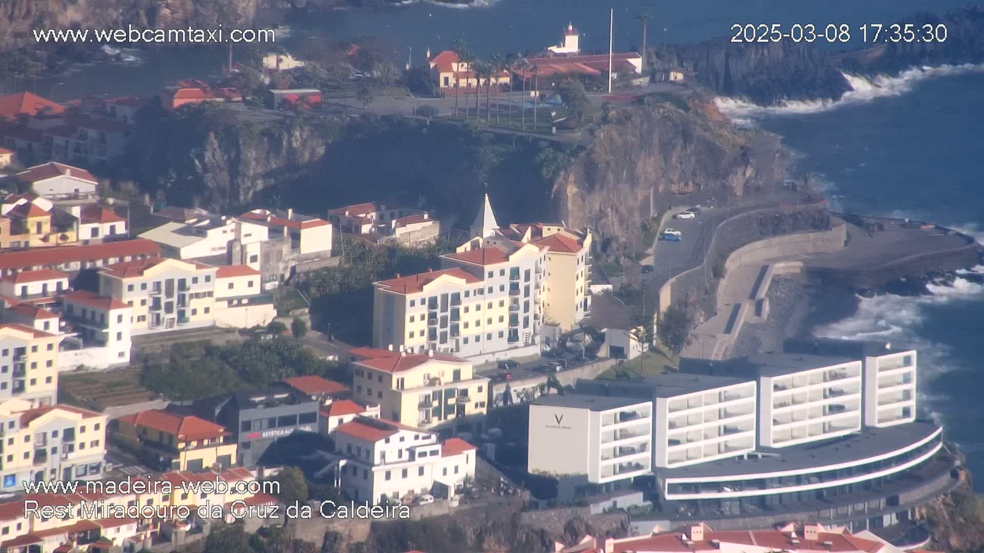 A coastal town with buildings of varying heights and styles sits on a cliff overlooking a rocky shoreline with crashing waves in sunny weather.