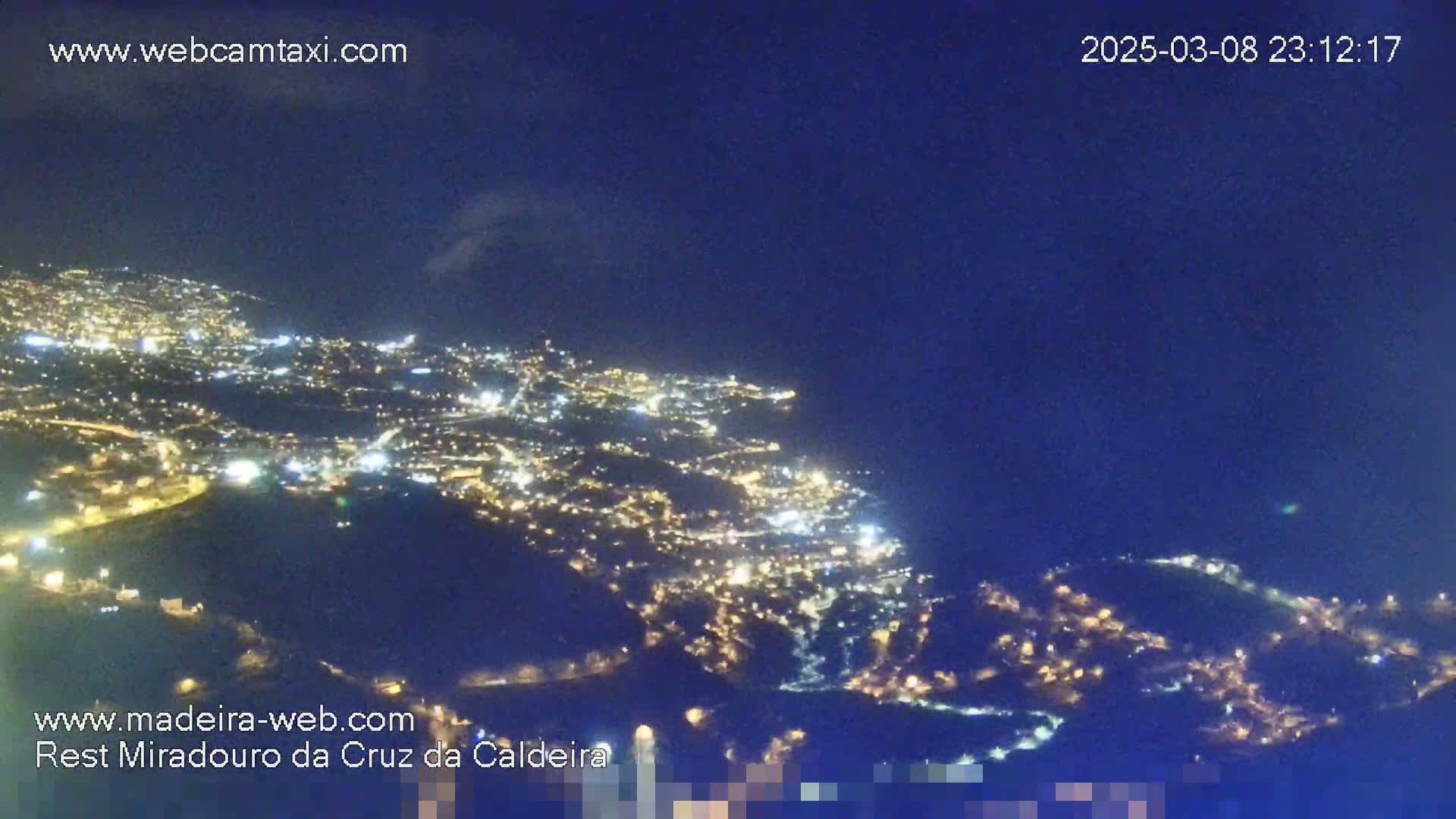 A nighttime, high-angle view shows a city illuminated with many lights spread across a mountainous landscape under a dark blue sky.