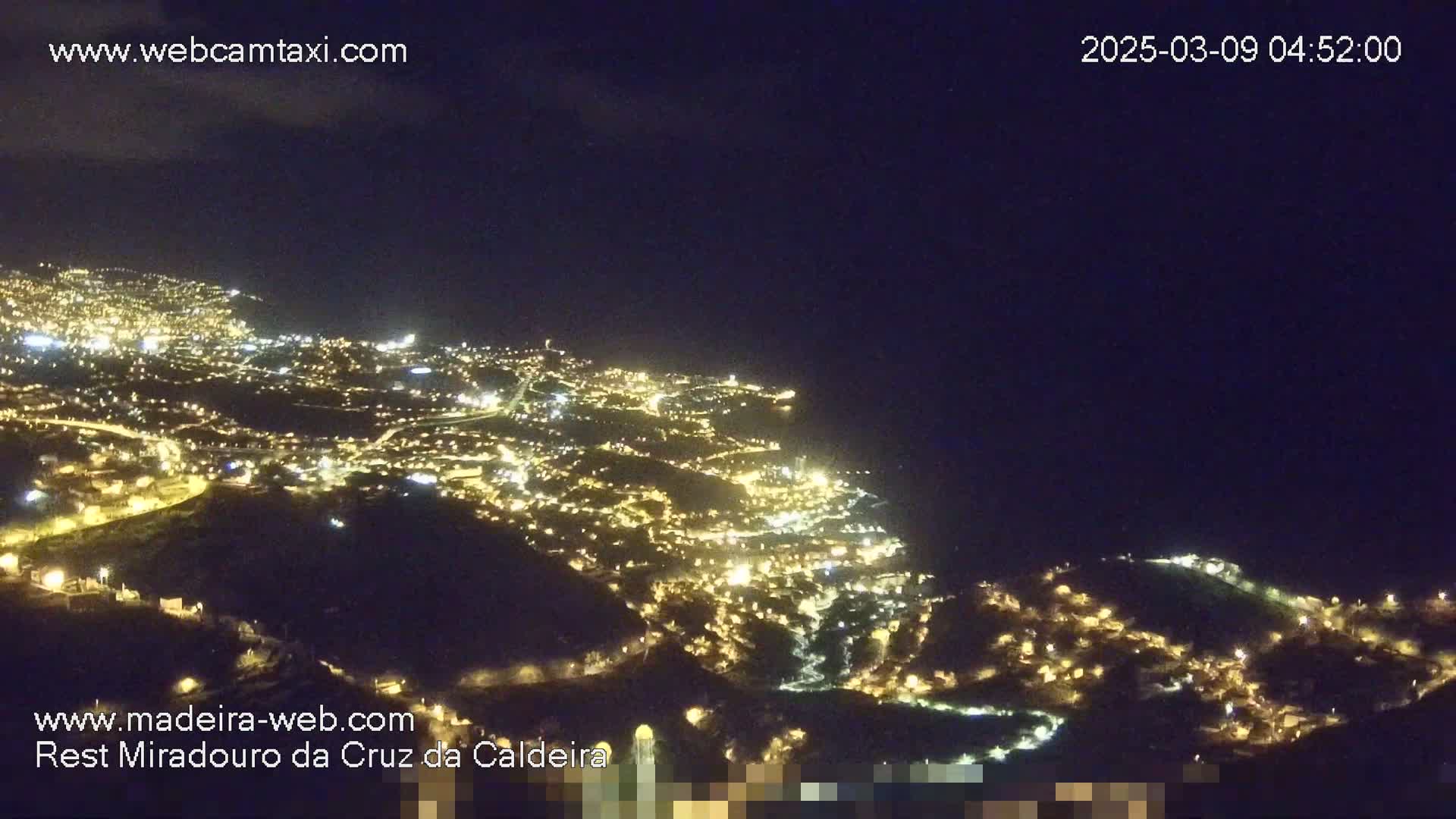 A nighttime, high-angle view shows a coastal town illuminated, with the dark ocean visible beyond.