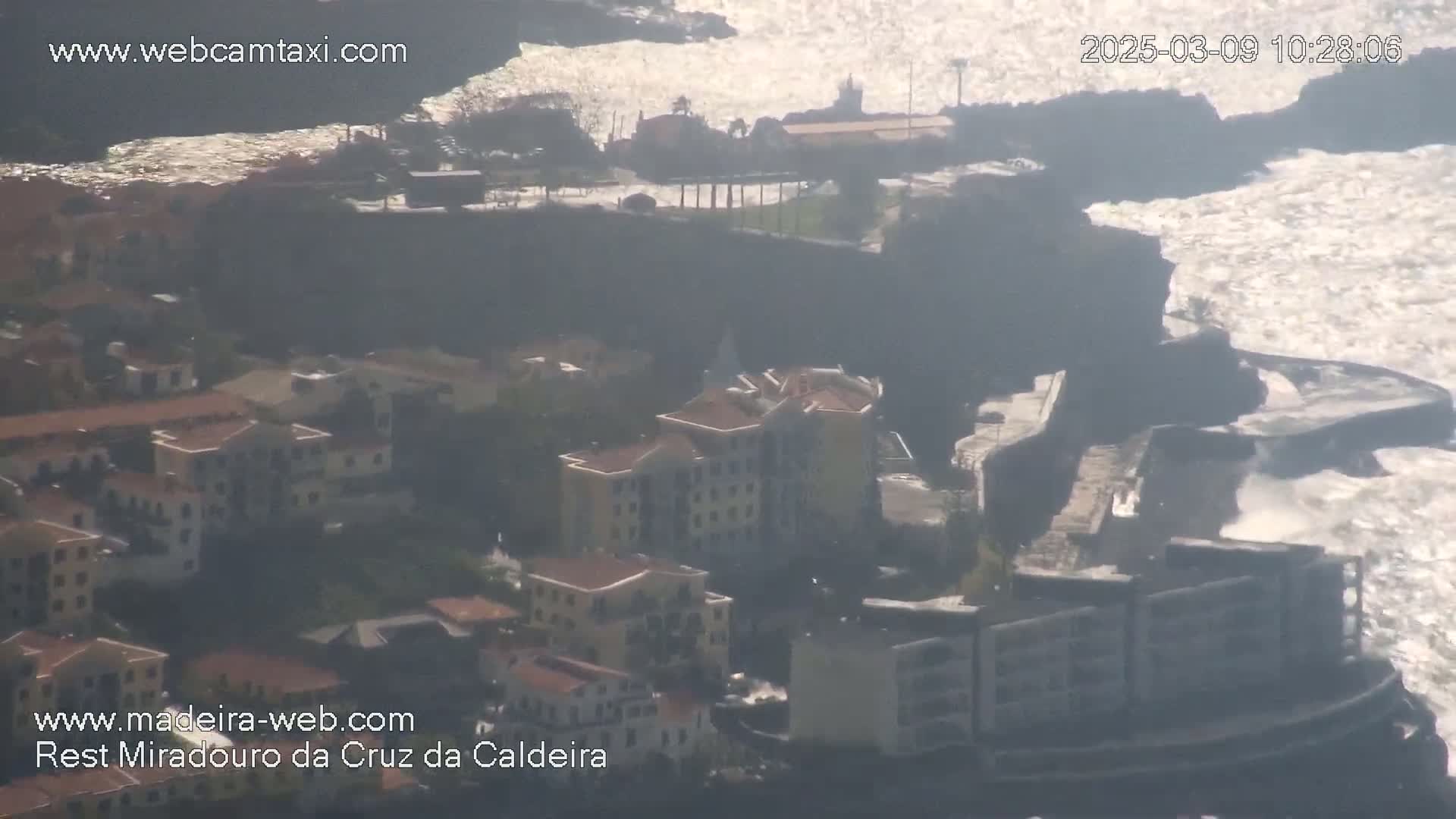 An aerial view shows a coastal town with buildings nestled against a rocky cliff face, where waves crash against the shore on a bright, somewhat hazy day.