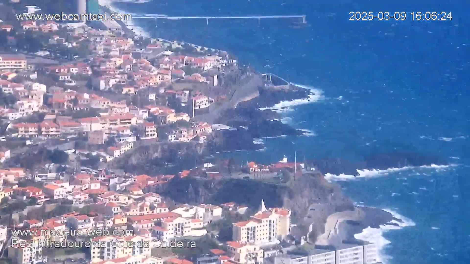 A coastal town built on cliffs overlooks a turbulent ocean with a bridge visible in the distance.
