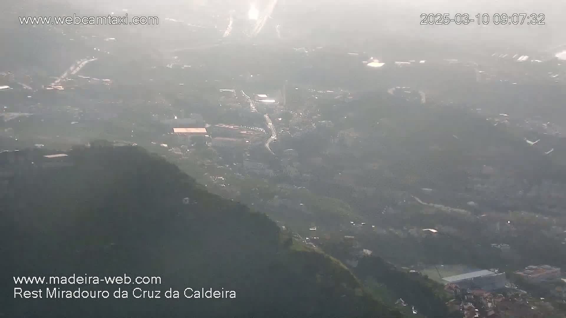 A hazy, high-angle view shows a valley with buildings and roads below a mountainside.