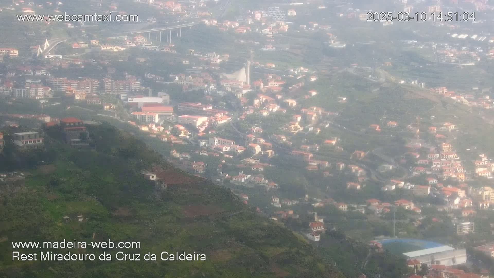 A hazy, high-angle view shows a hillside town with many small buildings, a bridge in the distance, and a large expanse of green hills.