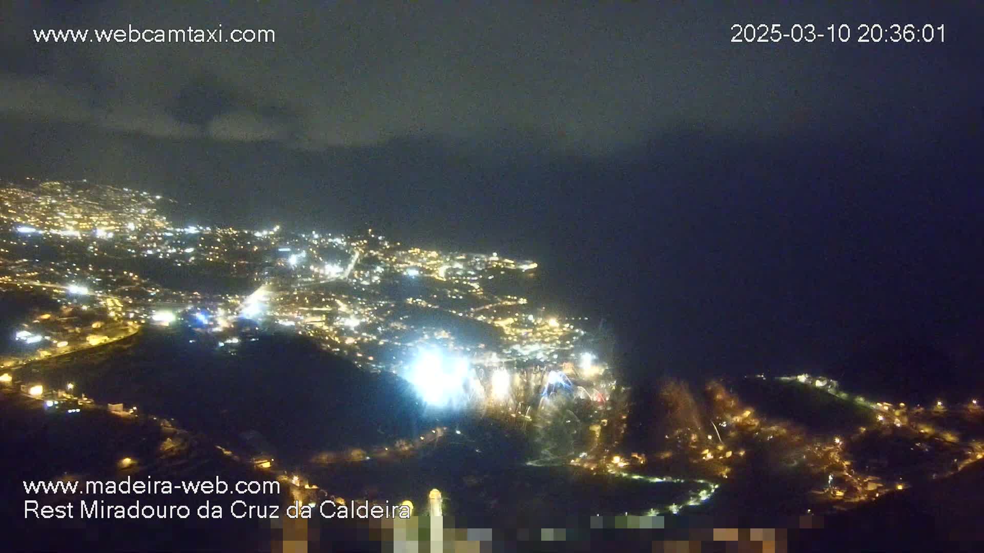A nighttime, high-angle view shows a coastal city illuminated with lights and a fireworks display, under a mostly cloudy sky.