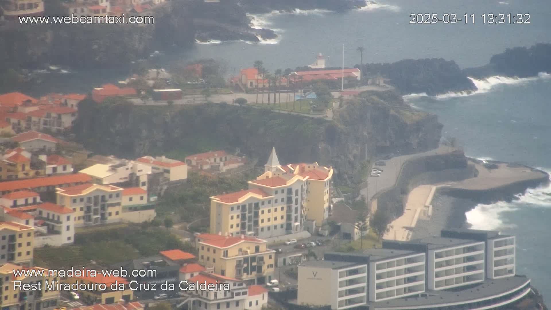 A coastal town with multi-story buildings is nestled against a rocky cliff overlooking a partially hazy ocean with waves crashing against the shore.