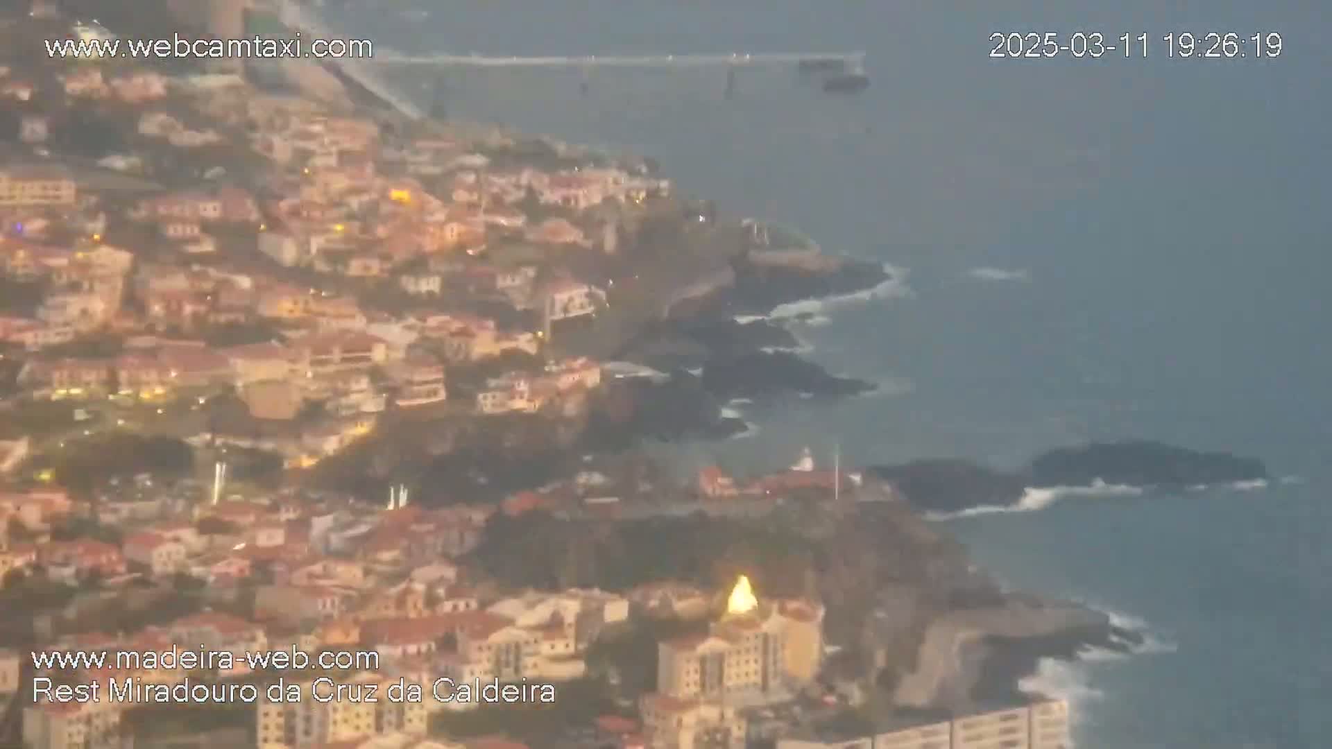 An aerial view shows a coastal town with many buildings illuminated at twilight, overlooking a rocky coastline with ocean waves under an overcast sky.