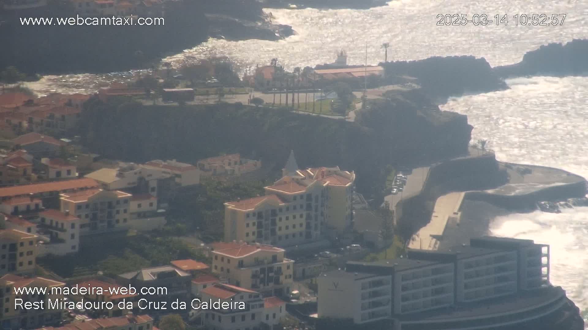 An aerial view shows a coastal town with buildings clustered on a cliff overlooking a rocky shoreline with moderately rough ocean waves.