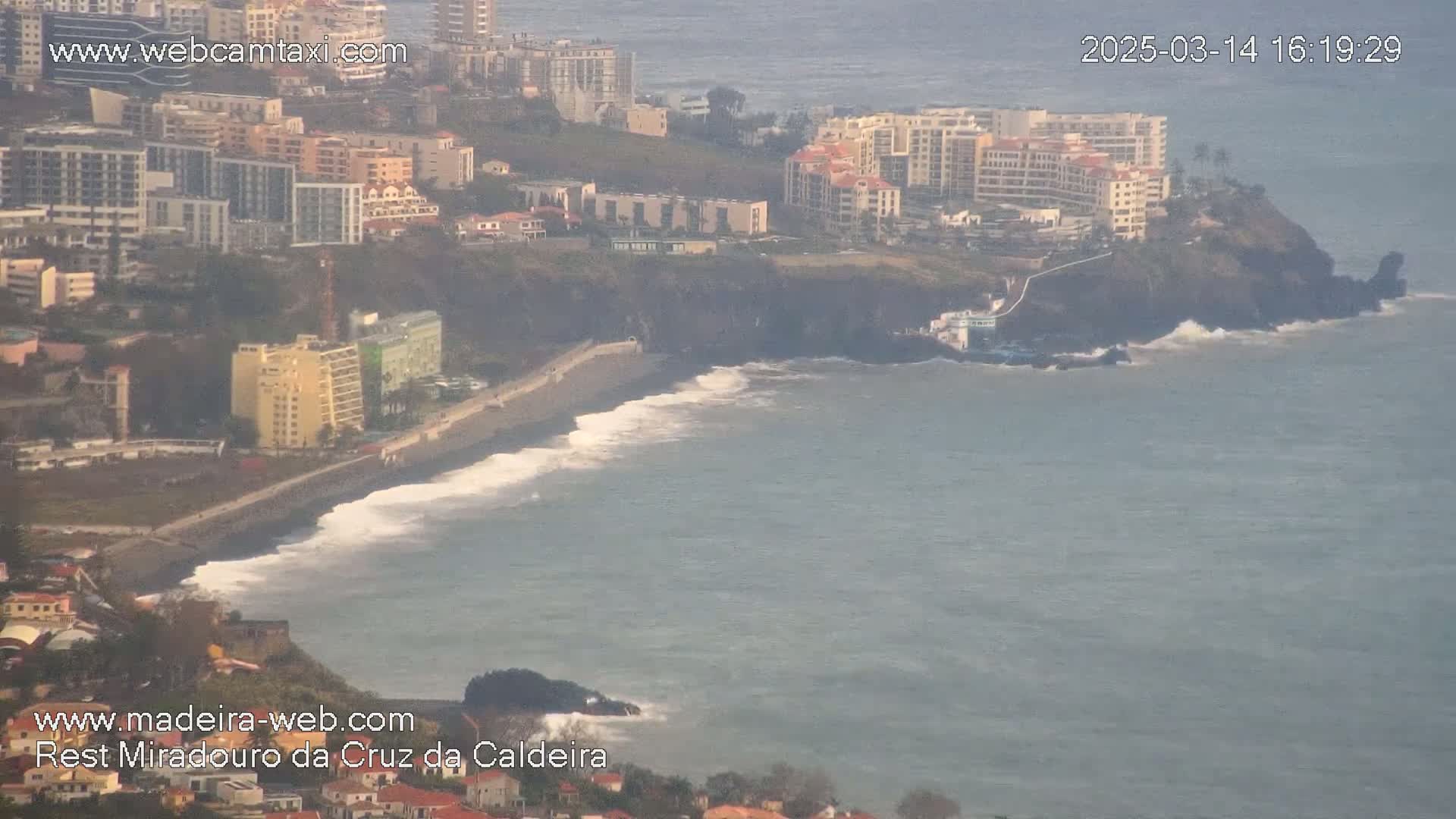 A coastal scene shows a dark-sand beach with moderate waves lapping the shore, buildings lining the cliffside, and hazy weather conditions.