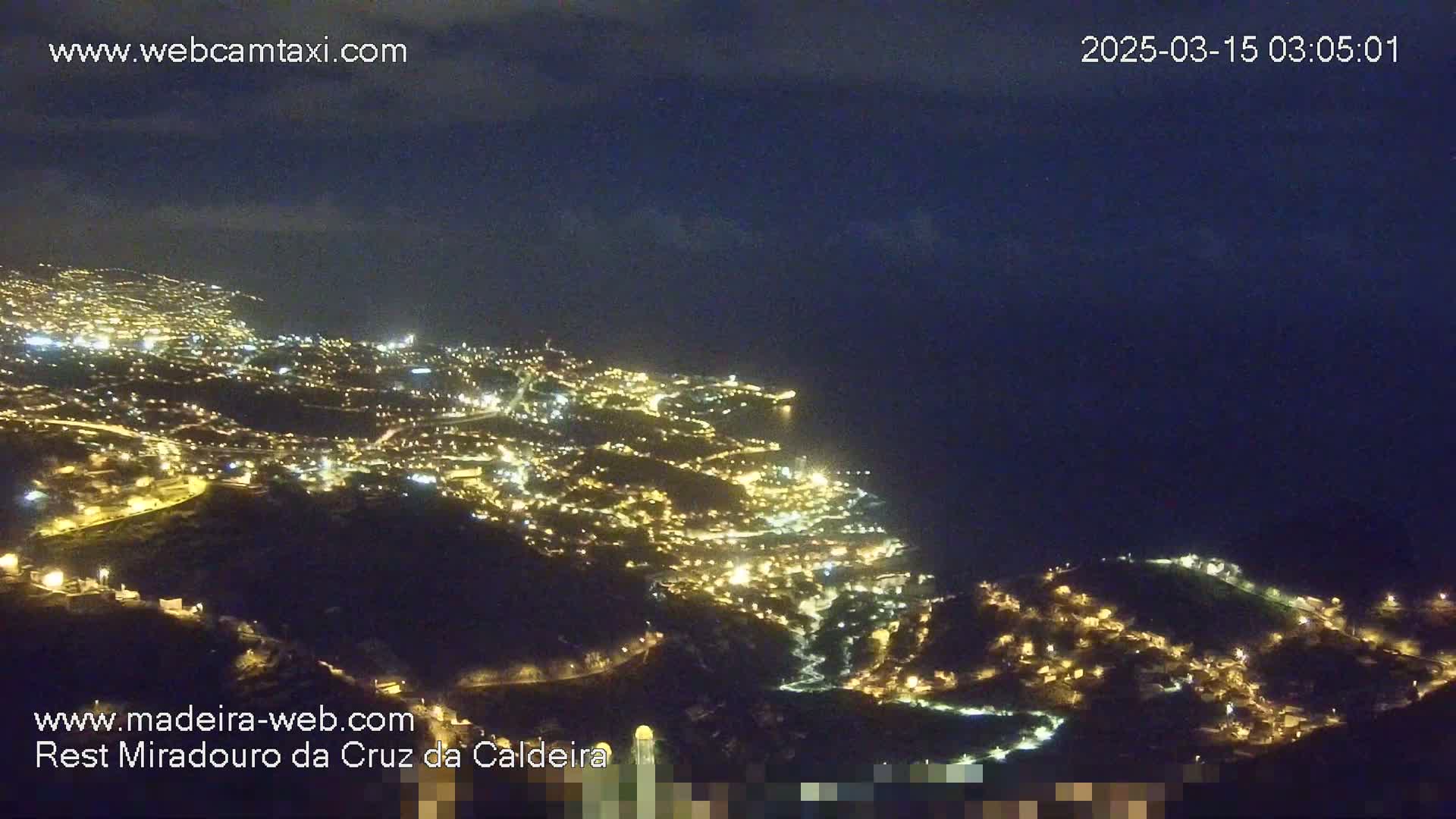 A nighttime, high-angle view shows a coastal city illuminated by many lights, extending down a hillside towards a dark ocean under a mostly clear night sky.