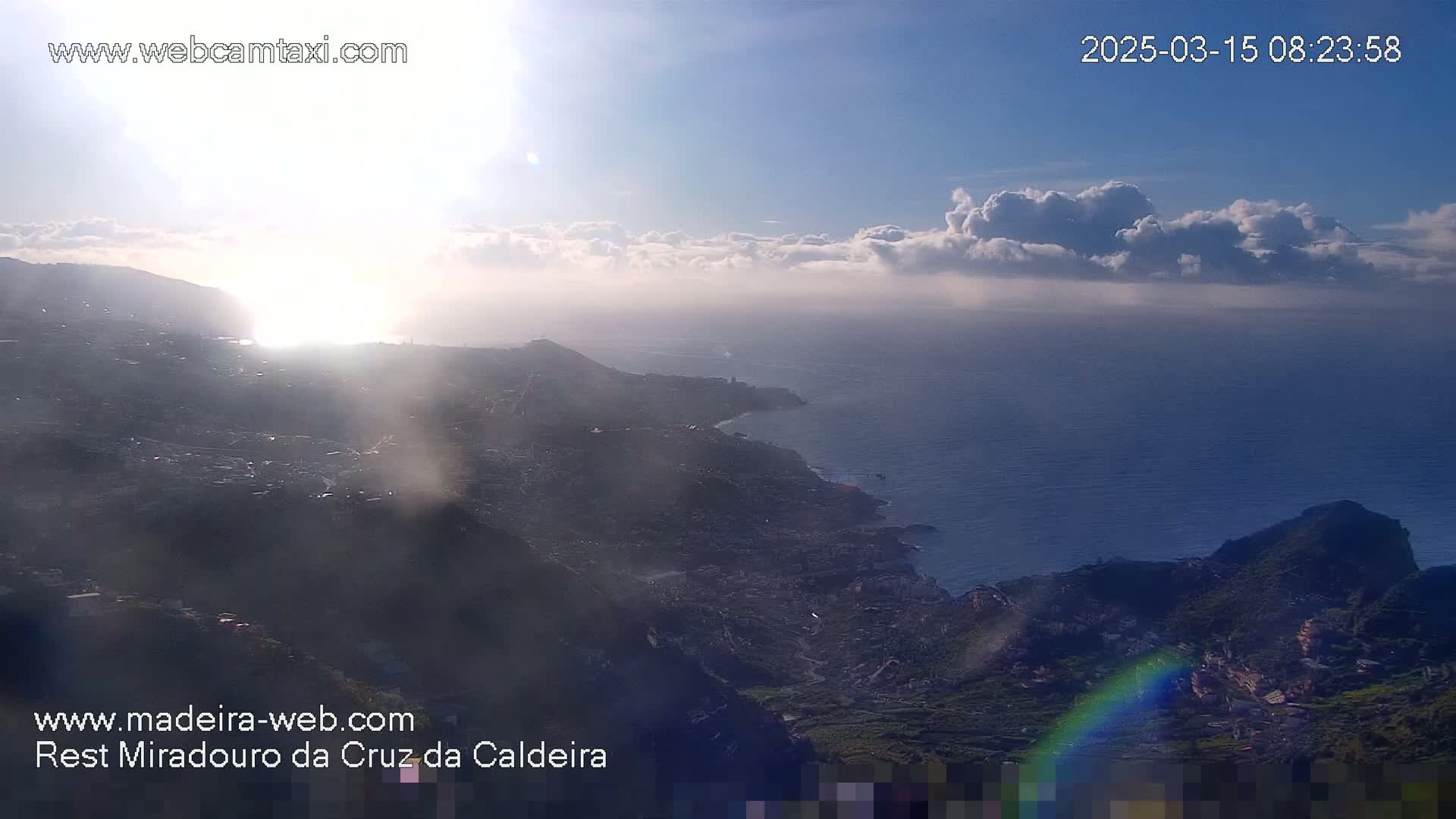 A coastal town nestled in a mountainous region is partially obscured by hazy sunlight and clouds, with a body of water visible in the background.