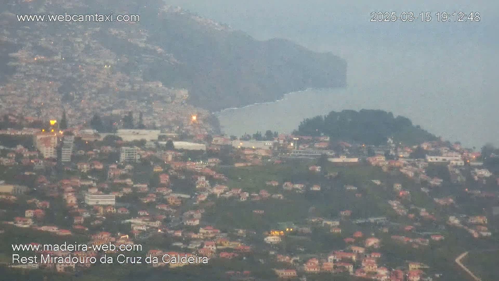 A hazy, dusk view shows a coastal town stretching across a hillside toward a calm ocean, with buildings and lights visible.