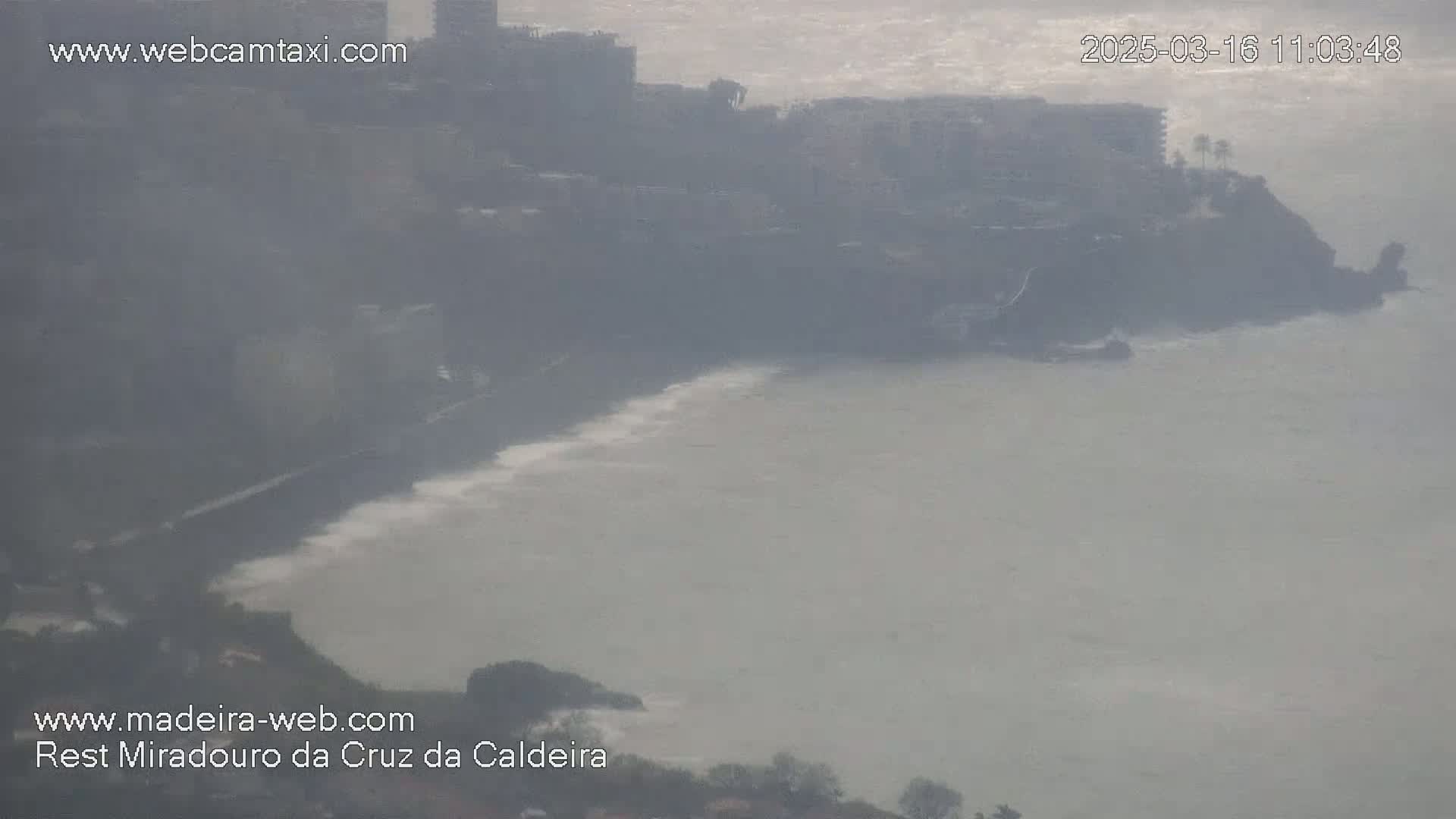 A hazy, overcast day reveals a coastal town nestled against a rocky shoreline with waves gently lapping at the shore.