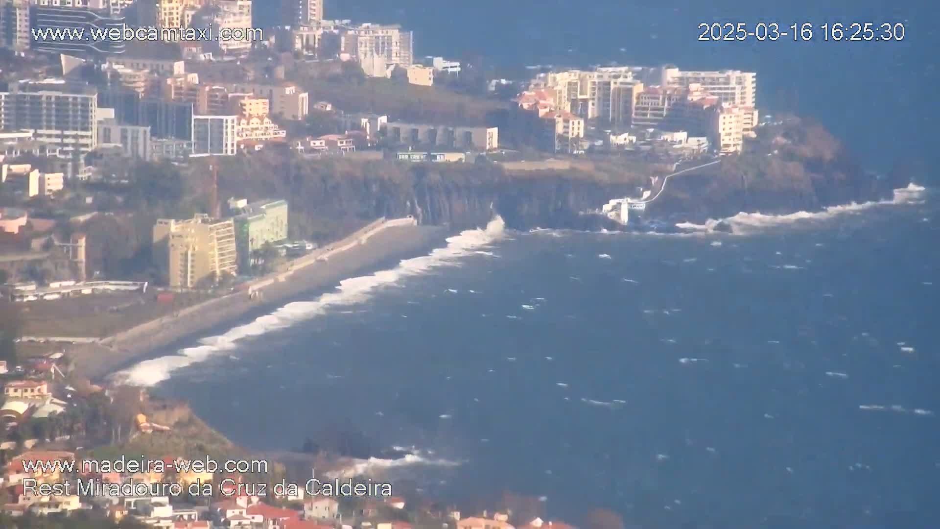 A coastal city with buildings and a beach is shown, with moderately rough ocean waves breaking on the shore under a clear sky.