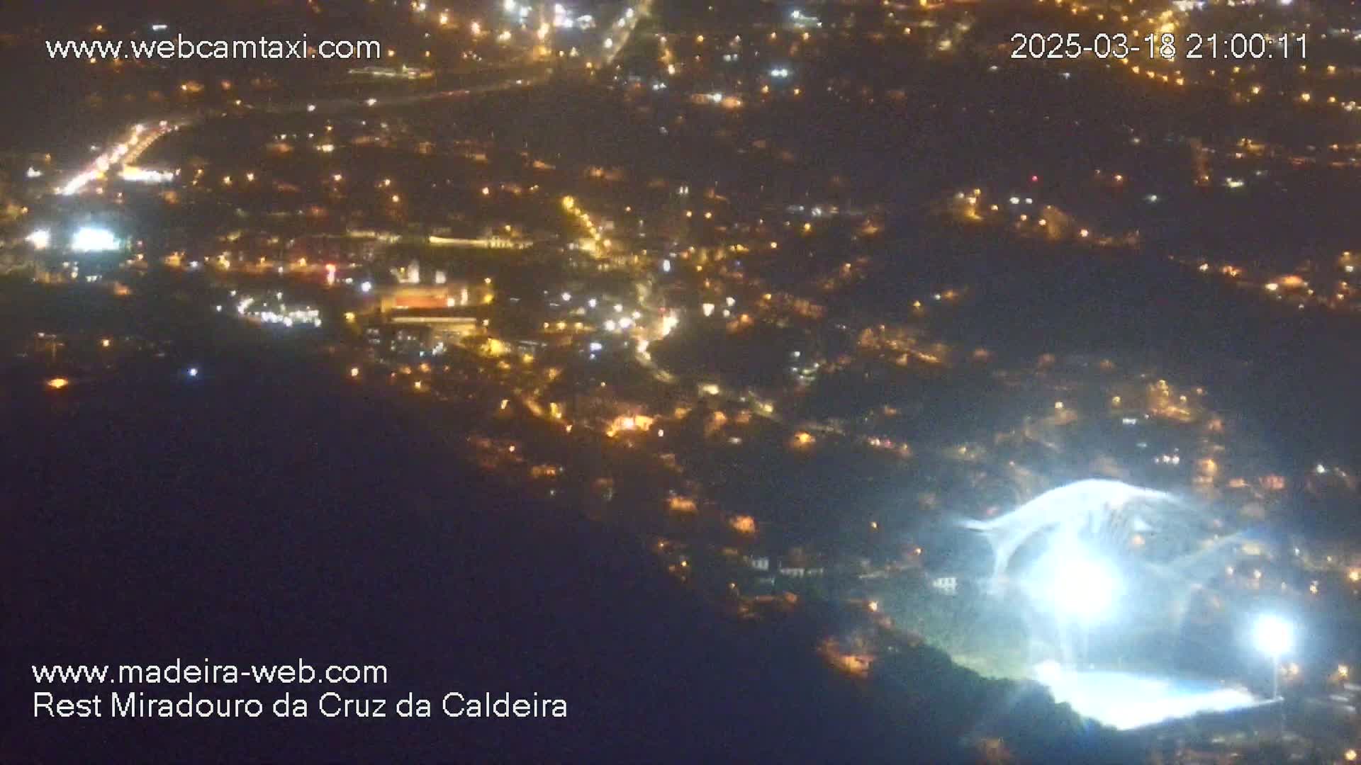 A nighttime, high-angle view shows a brightly lit area, possibly a stadium, below a city illuminated with scattered lights.