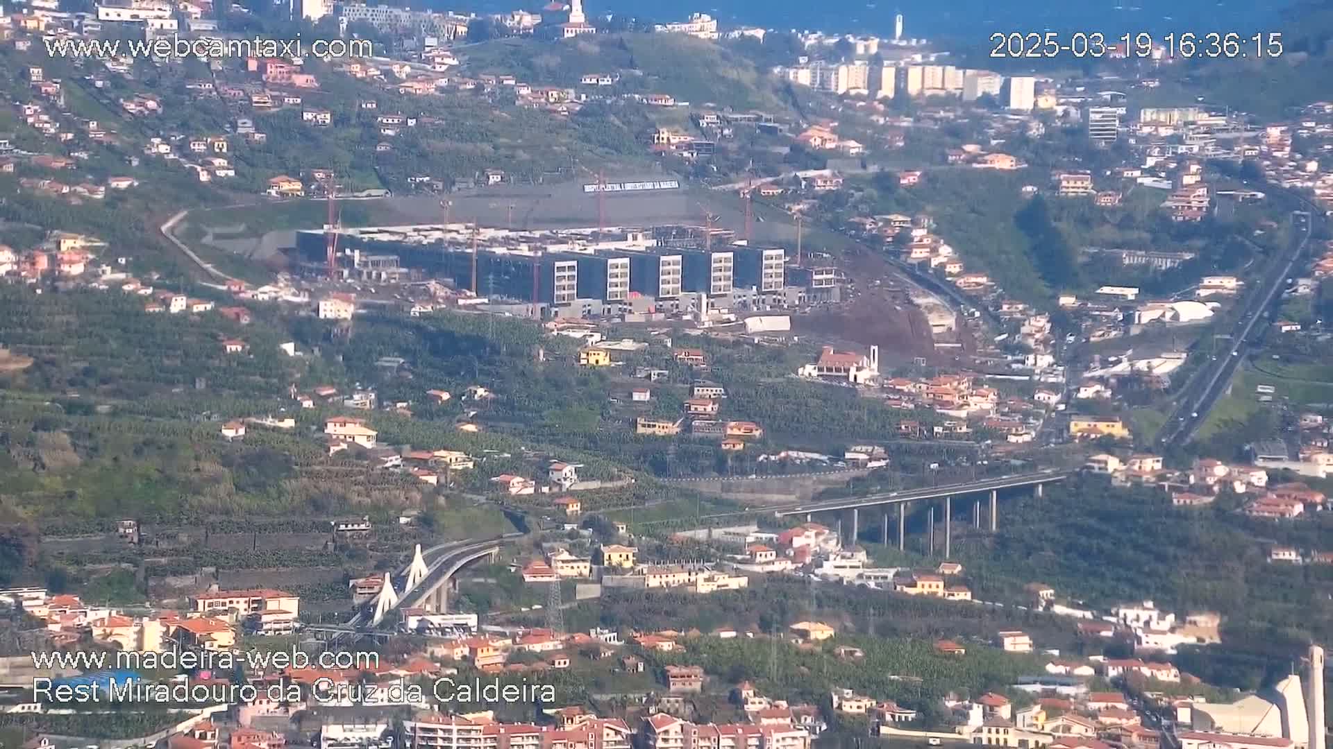A sprawling hillside town with a large building under construction, a bridge, and a highway is visible on a clear day.