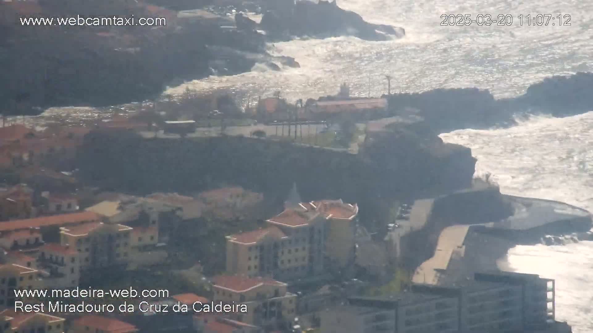 A coastal town with buildings clustered together is situated on a rocky cliff overlooking a turbulent ocean under a bright, sunny sky.