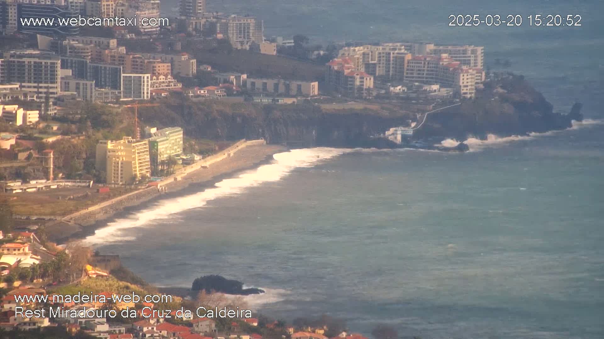 A coastal town with buildings and a black sand beach, where moderate waves are breaking onto the shore on a partly sunny day.