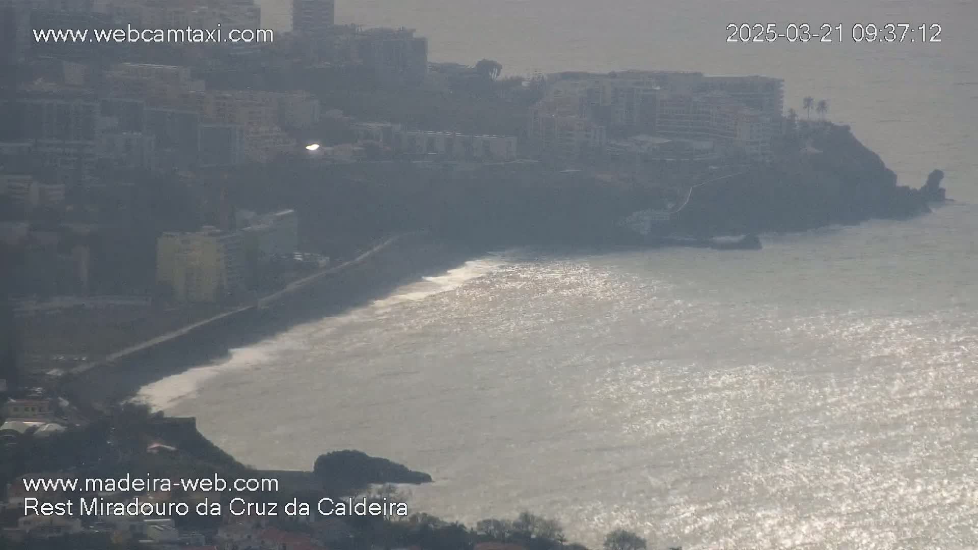A hazy coastal scene shows a rocky shoreline with buildings perched on a cliff overlooking a wavy ocean reflecting sunlight.