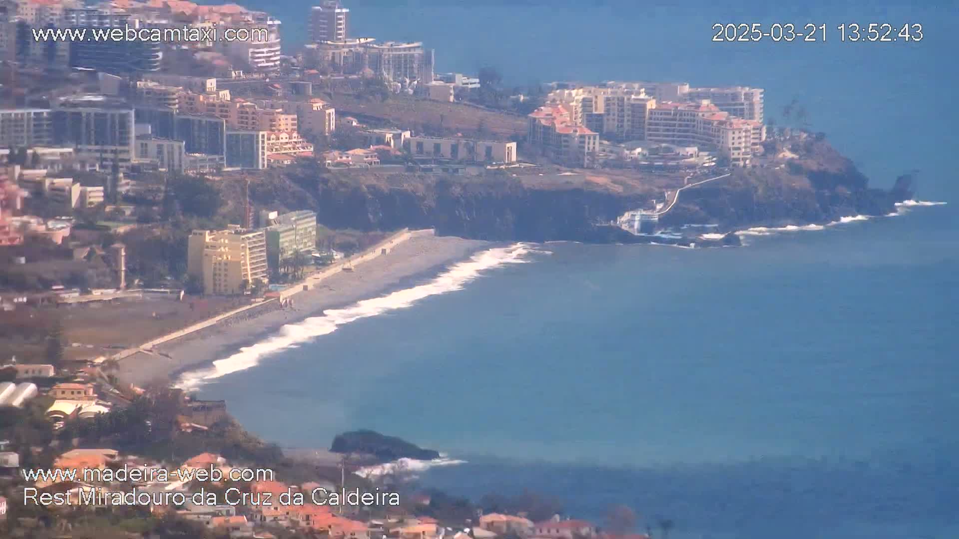 A coastal town with buildings on a cliff overlooking a dark-sand beach and a blue ocean with small waves under a sunny sky.