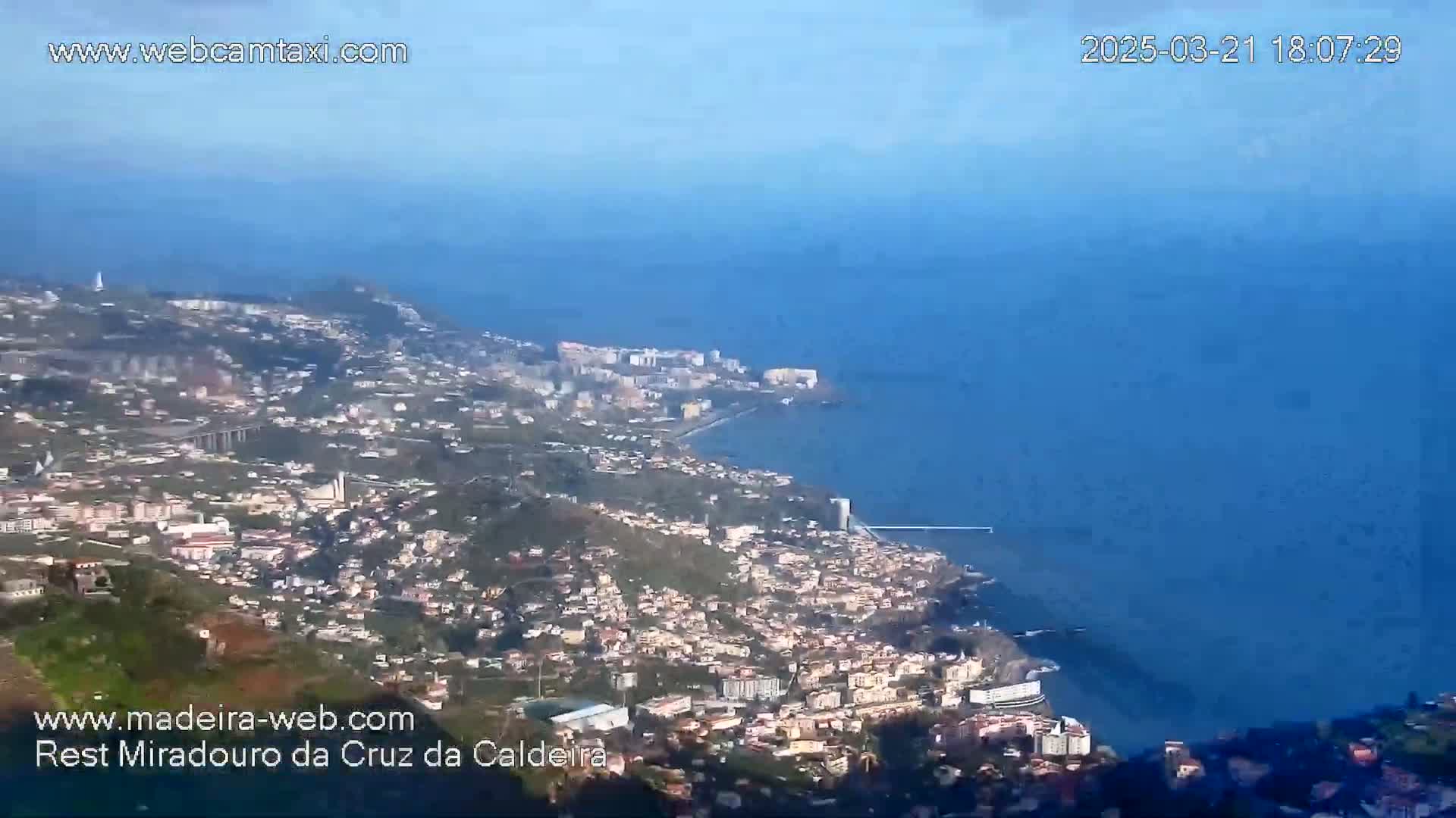 A coastal town sprawls along a hillside overlooking a calm ocean under a clear sky.
