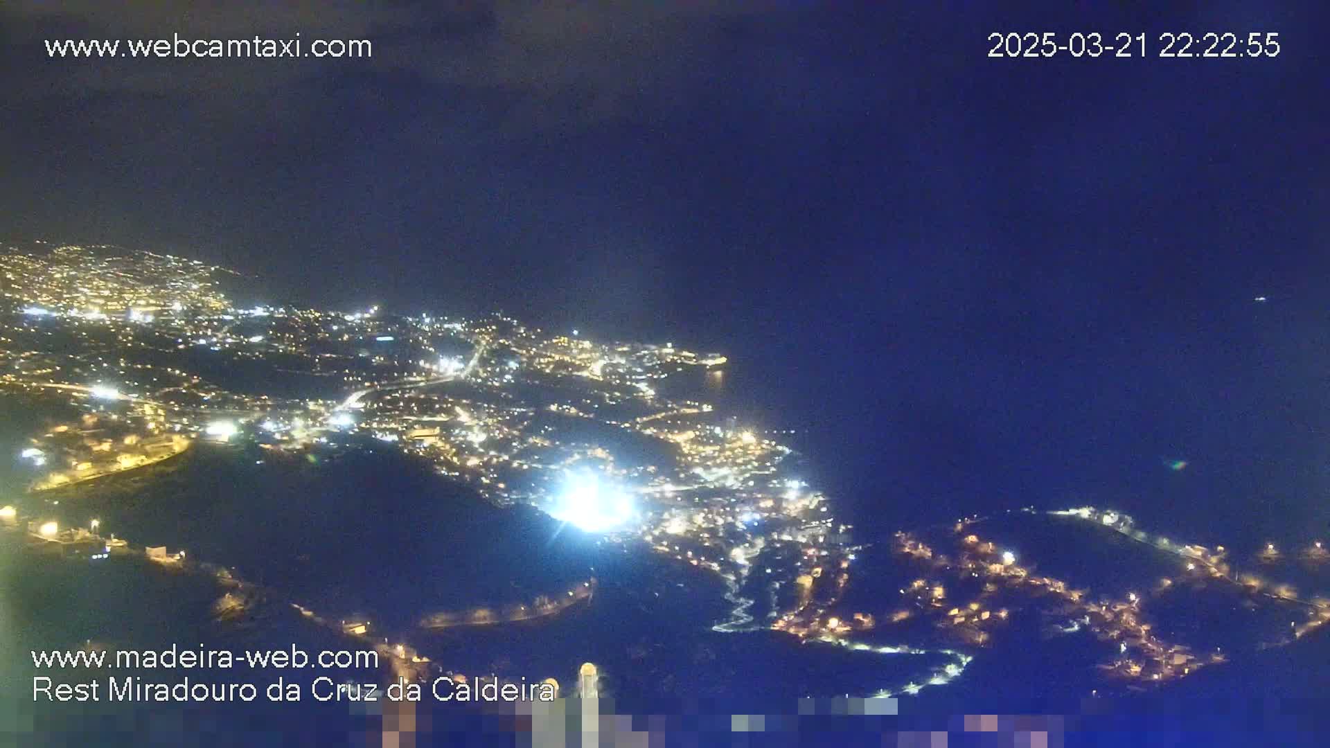 A nighttime view of a city illuminated by lights, next to a dark body of water under a clear, dark sky.