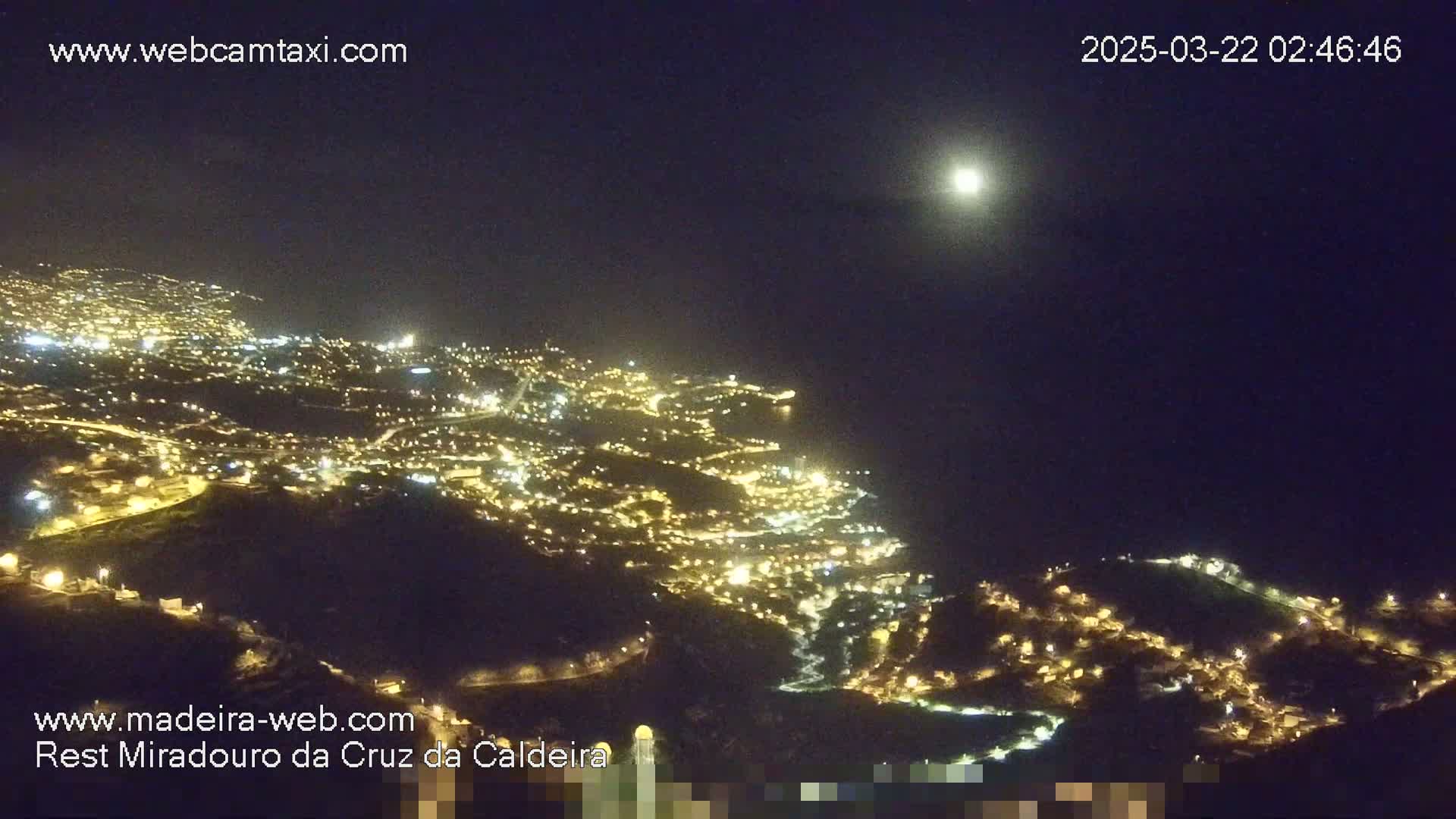 A nighttime aerial view shows a brightly lit city nestled on a hillside overlooking the dark ocean under a clear, moonlit sky.