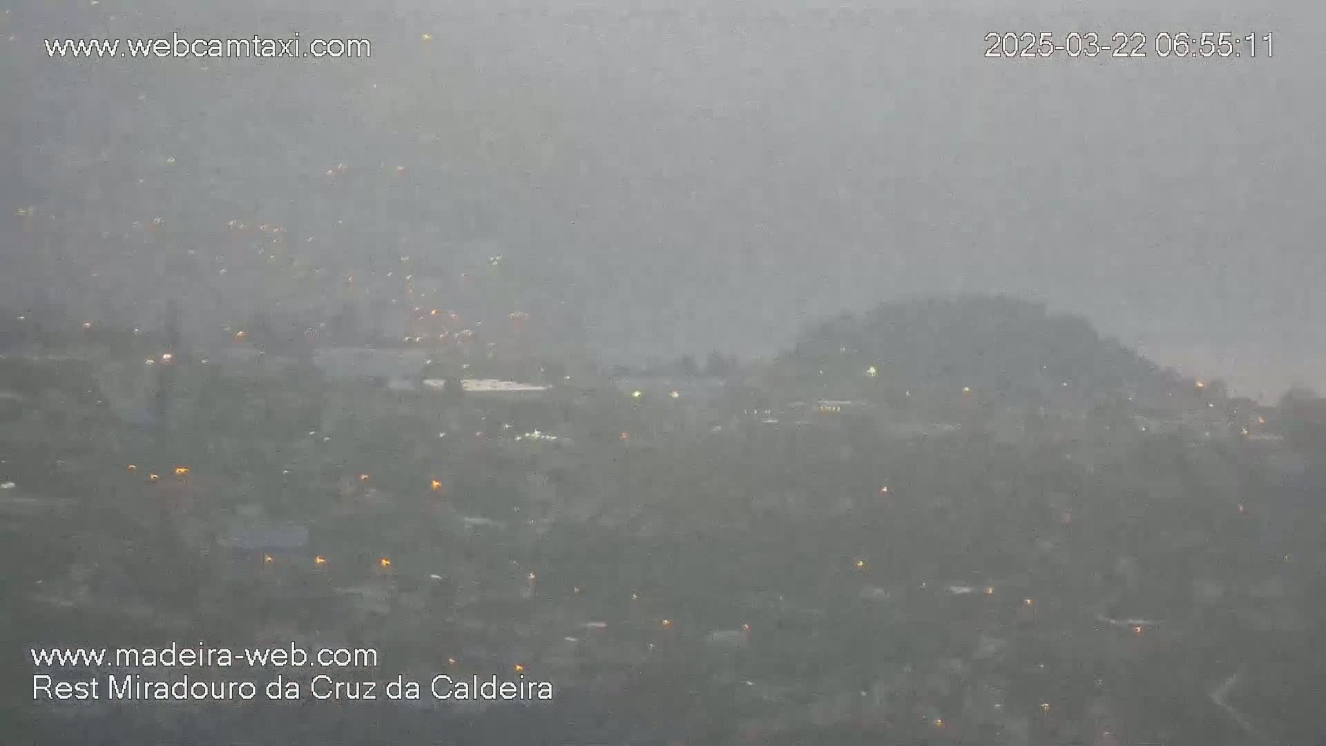 A hazy, overcast view of a hilly town with scattered lights visible through the heavy precipitation.