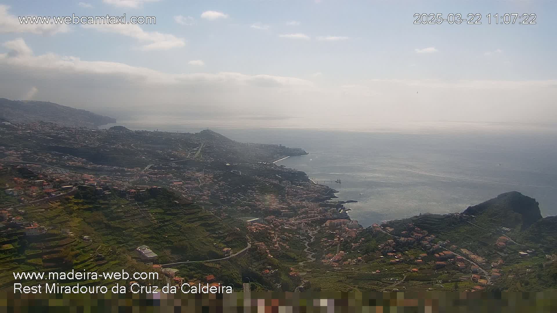 A coastal town nestled on terraced hillsides overlooking a calm ocean under a partly cloudy sky.