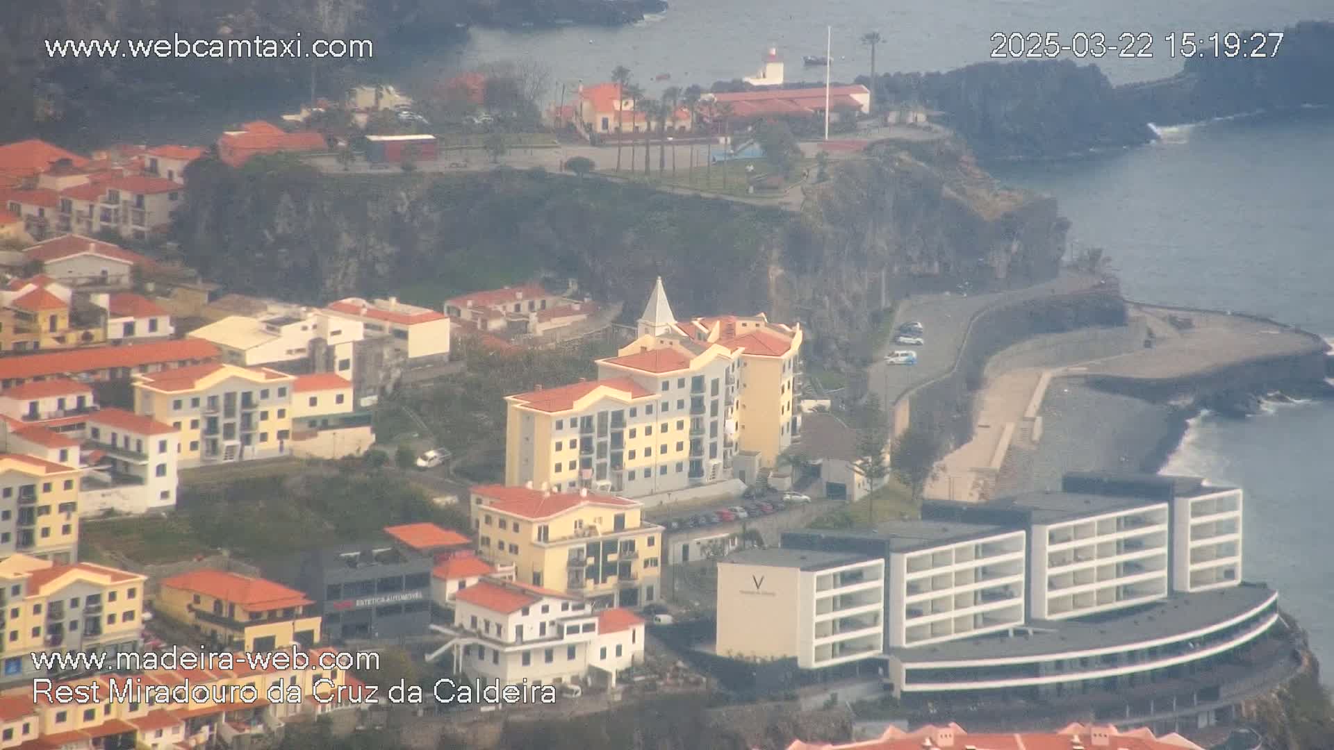 An aerial view shows a coastal town with multi-story buildings, a rocky shoreline, and a hazy, overcast sky.