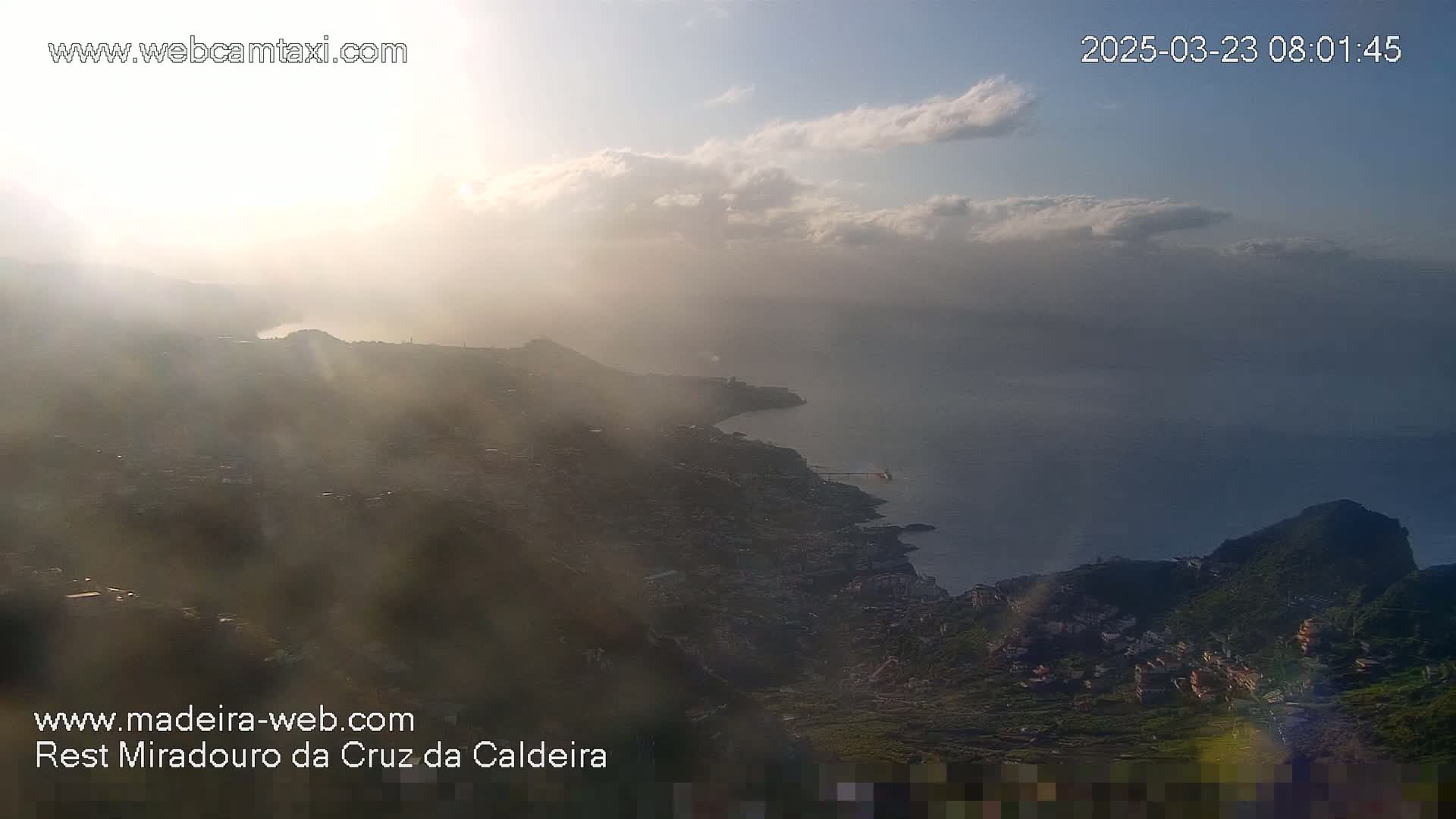 A hazy sunrise illuminates a coastal town nestled between hills and the ocean, with some clouds in the sky.