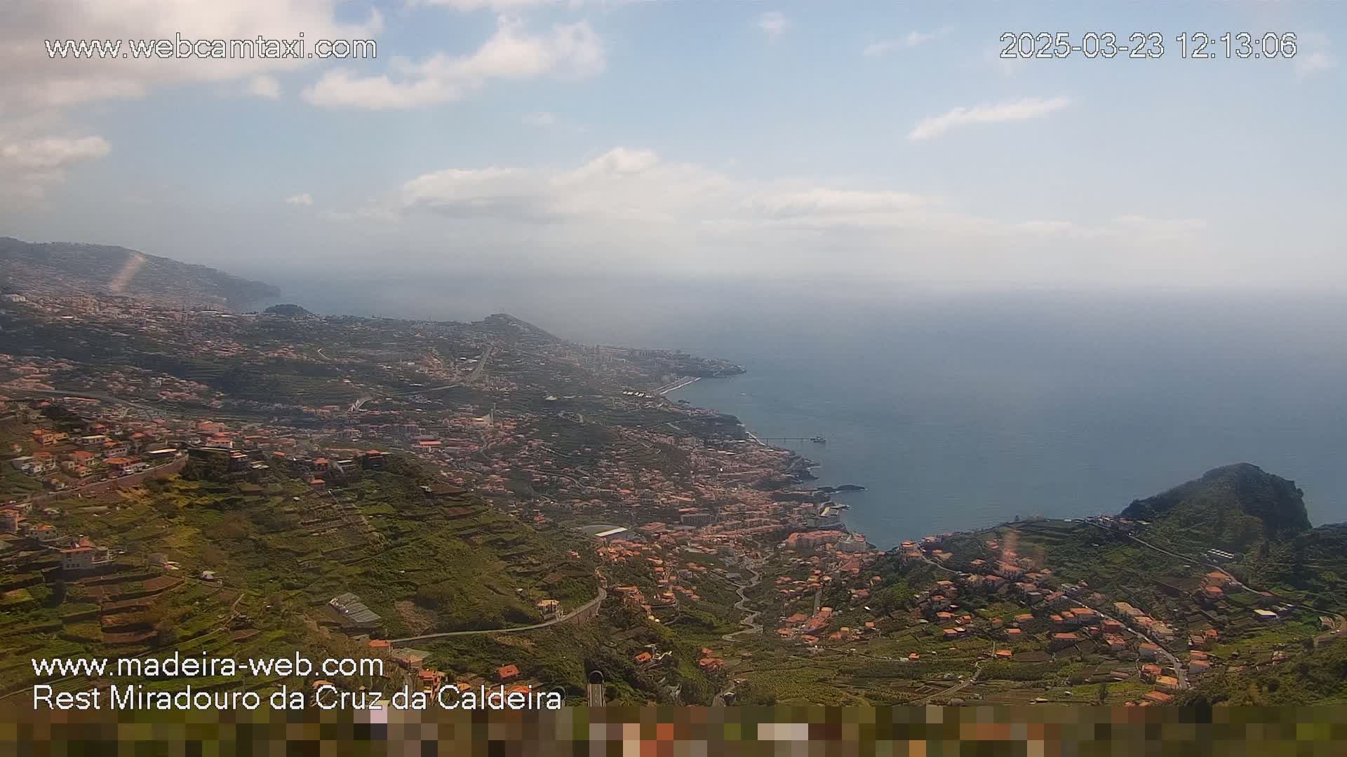 A coastal town nestled on a hillside overlooking a calm ocean under a partly cloudy sky.