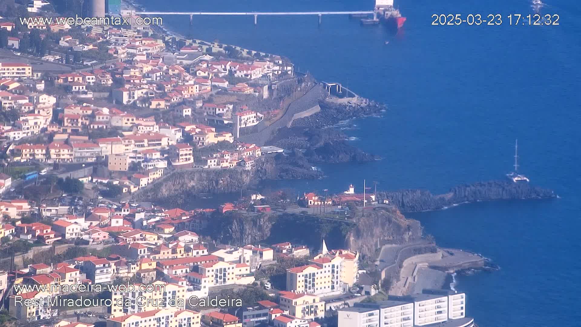 A coastal town with red-roofed buildings is situated on a cliff overlooking a dark blue ocean on a sunny day.